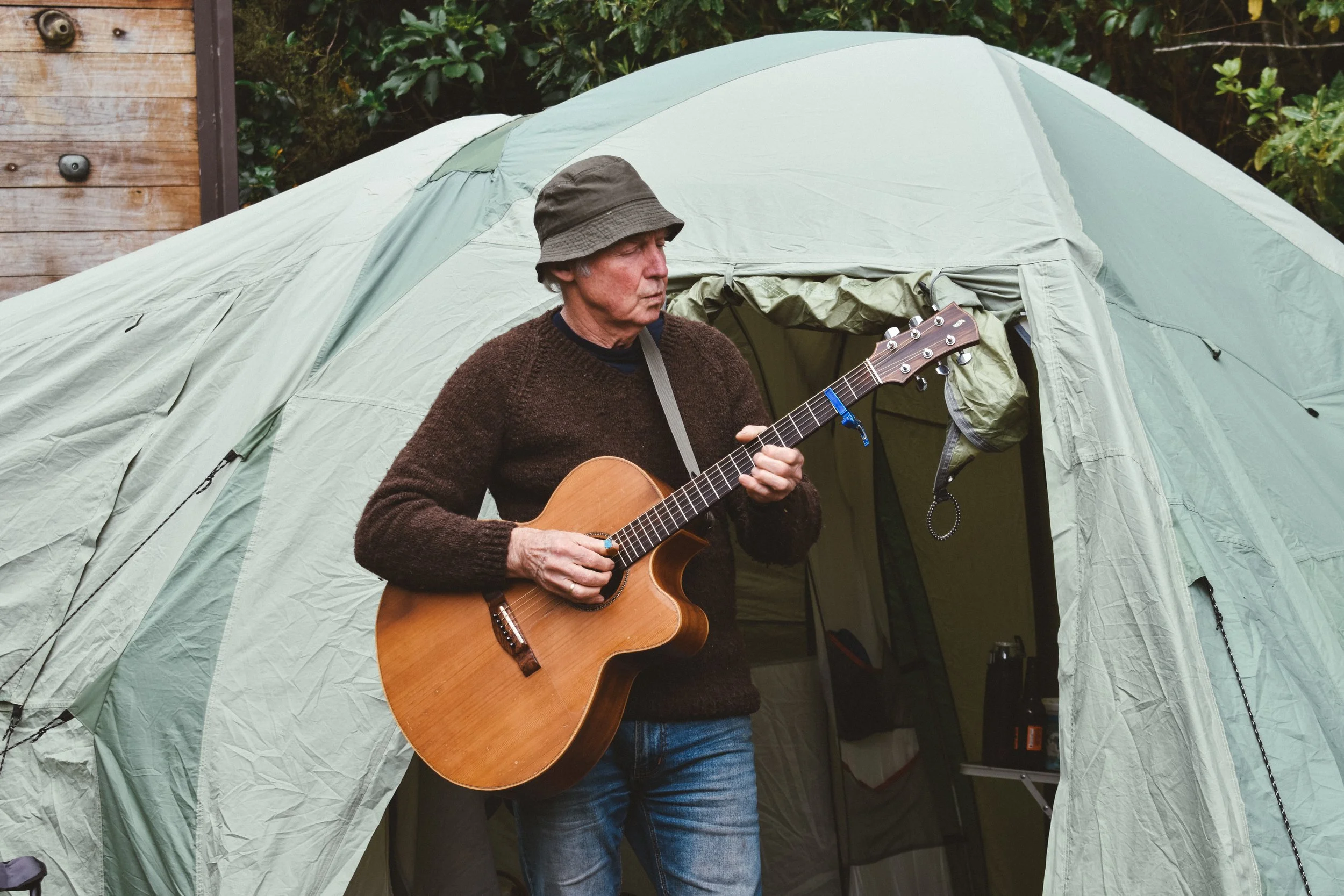 An older man wearing a brown sweater and a gray bucket hat playing an acoustic guitar outside a camping tent.