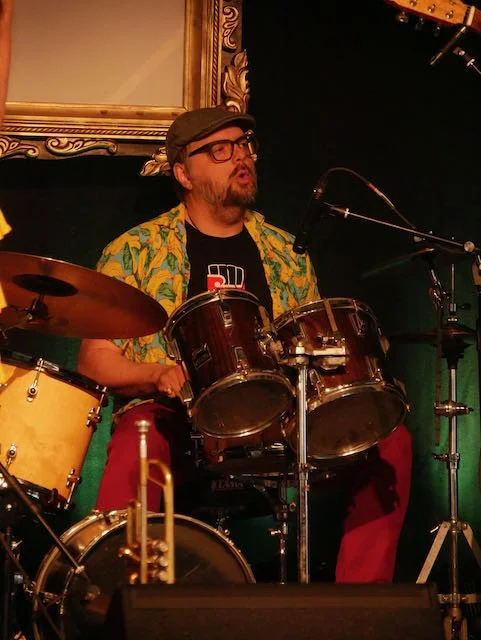 A man in a yellow floral shirt, black hat, and glasses playing drums and singing into a microphone on stage.