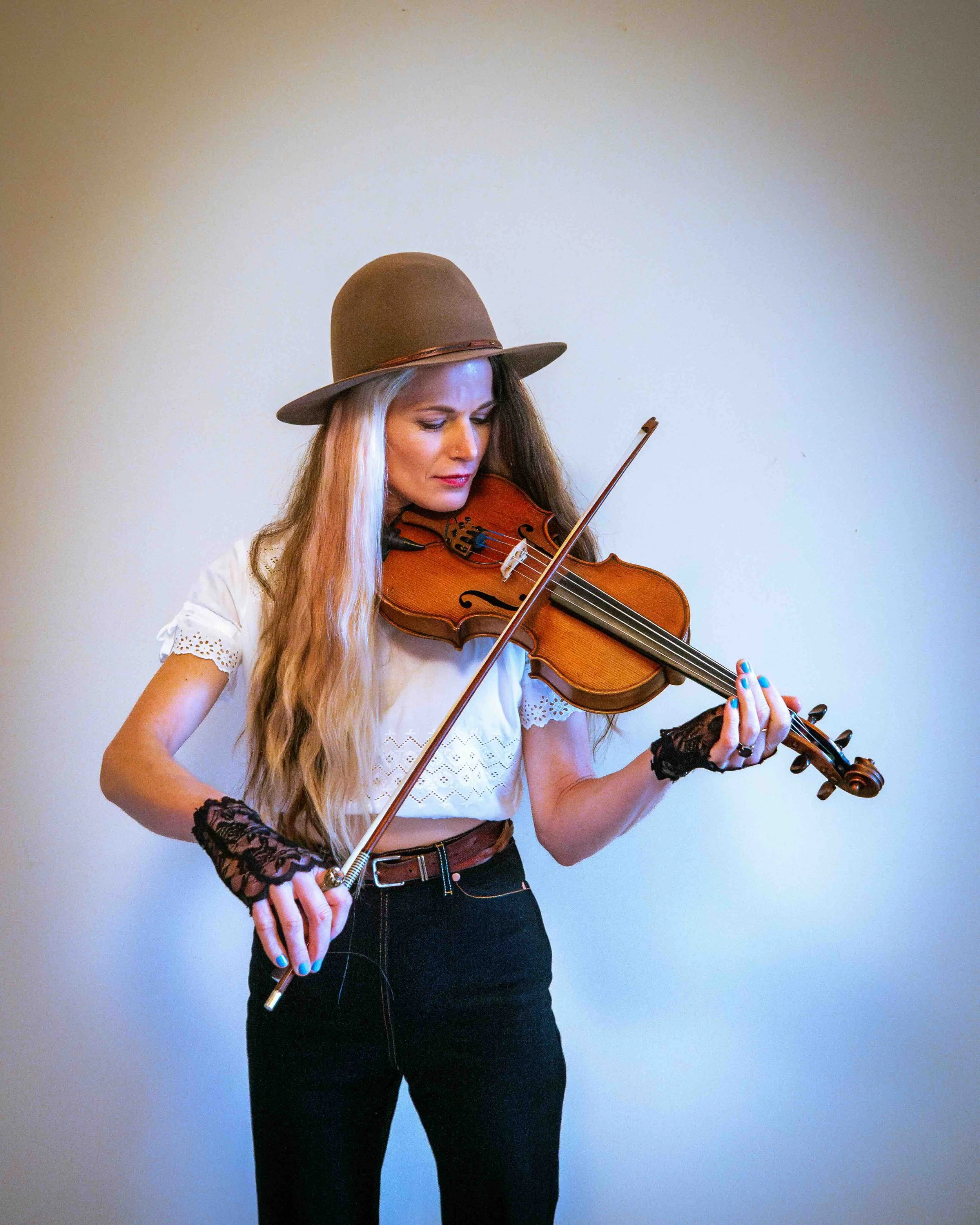 A woman with long blonde hair wearing a brown hat, white blouse, and black pants, playing the violin.