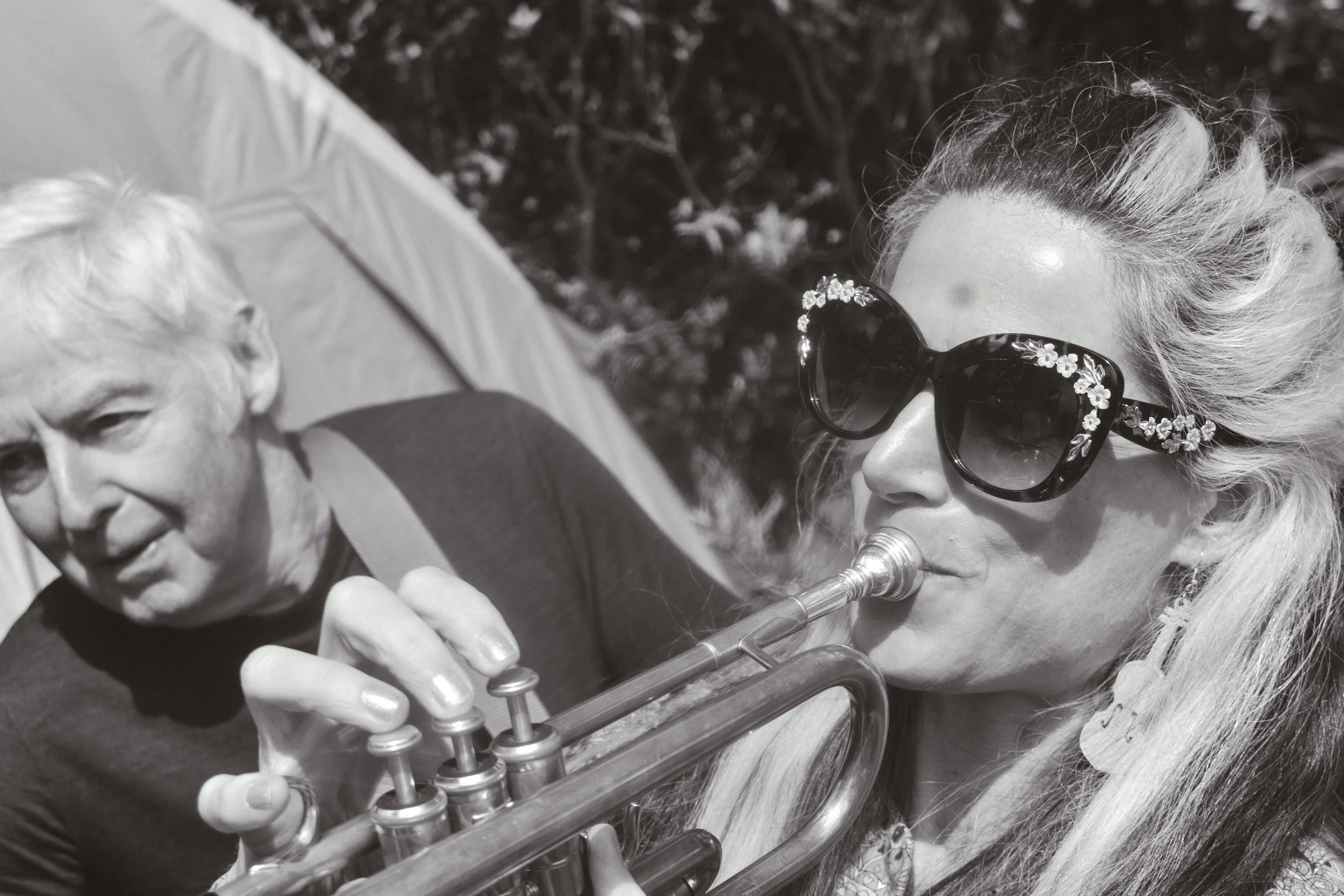 Woman wearing sunglasses playing trumpet outdoors, with older man observing.