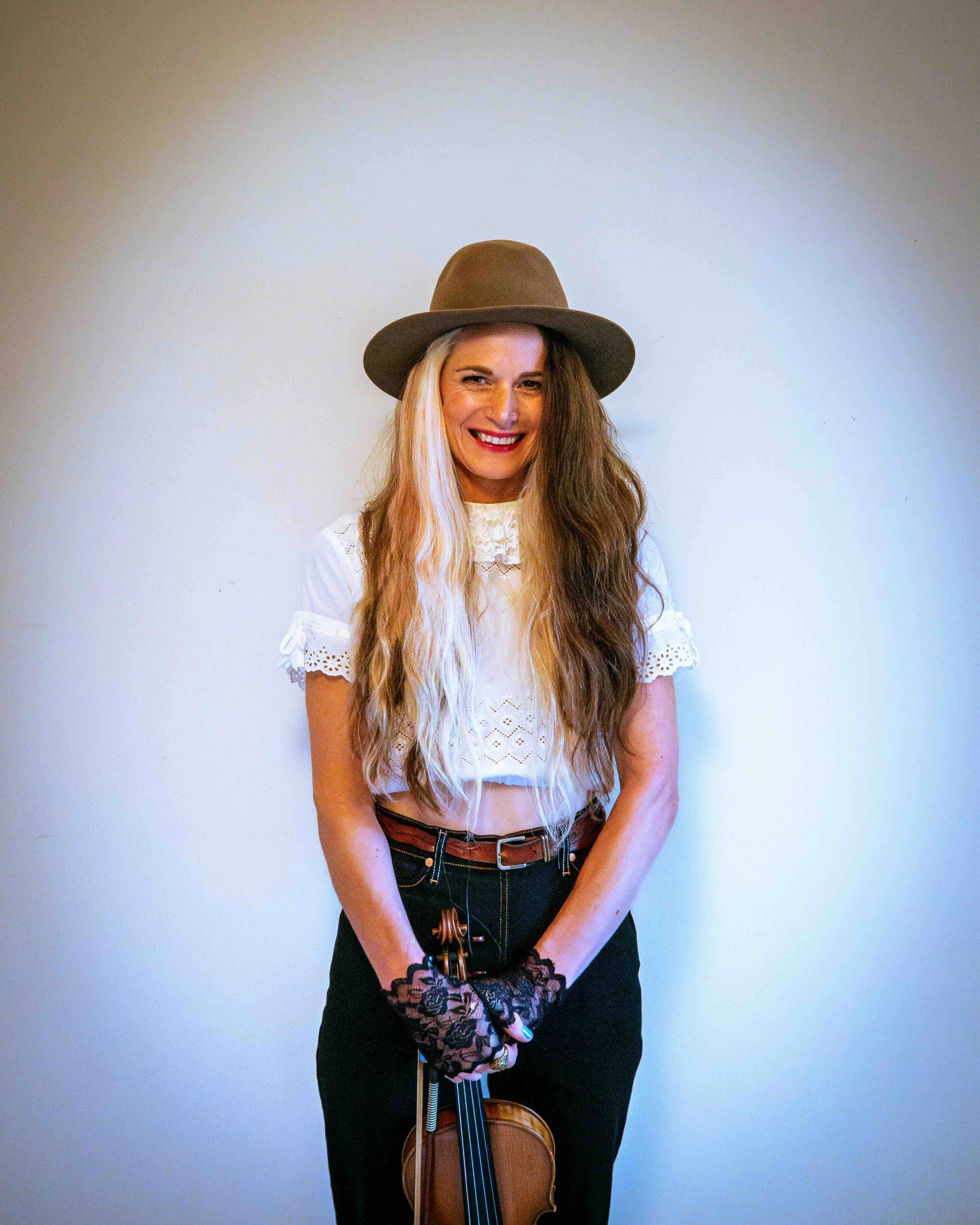Woman with long blonde hair wearing a brown hat, white eyelet blouse, black pants, and lace gloves, holding a violin against a plain wall.