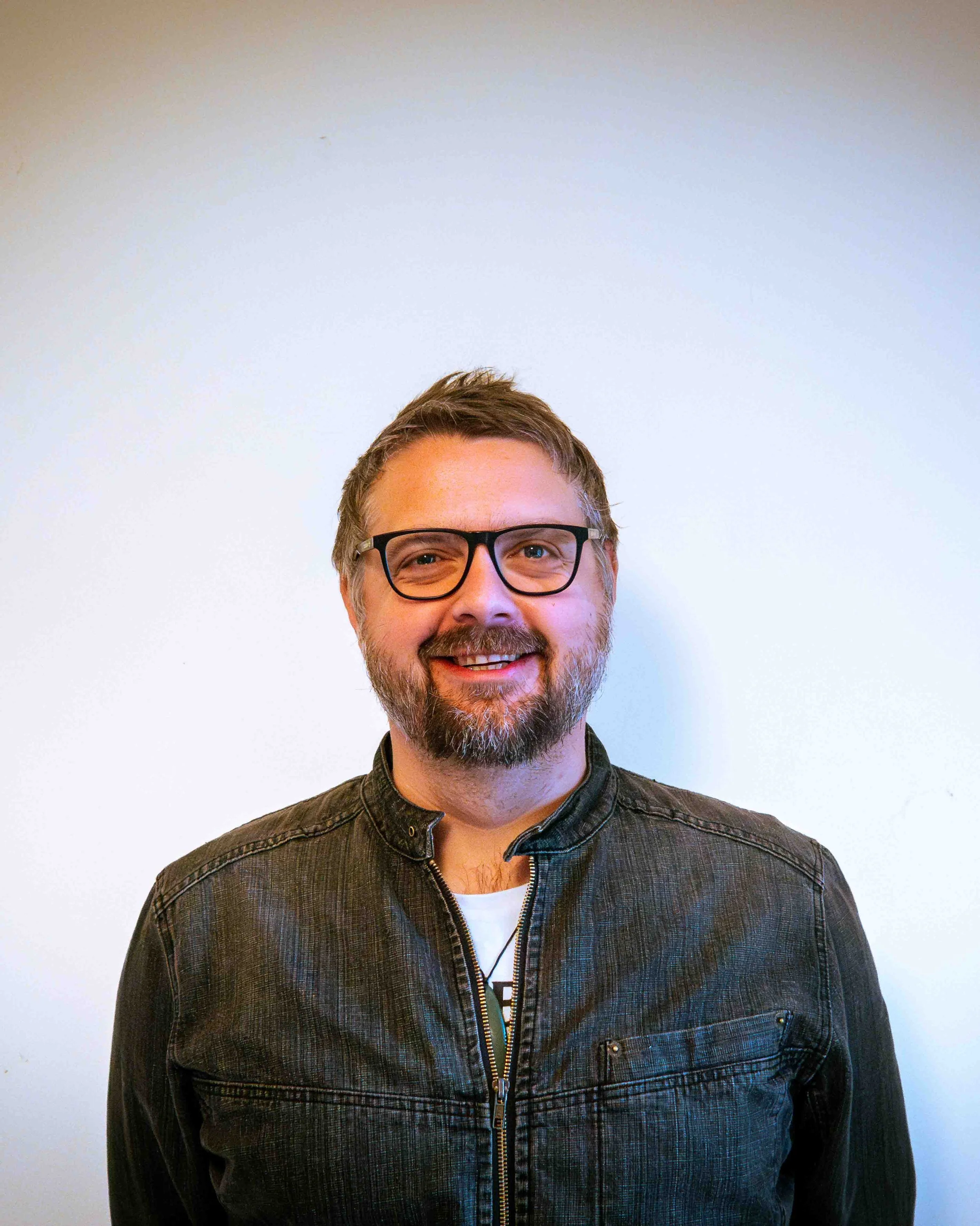 Smiling man with glasses, beard, and brown hair, wearing a black leather jacket, standing against a plain white wall.