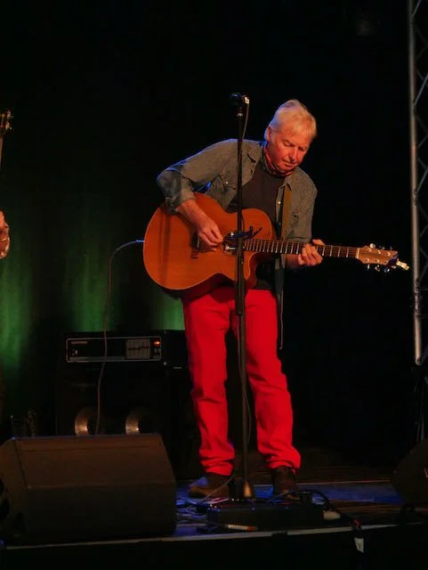 Older man playing acoustic guitar on stage with black background, microphone stand, and sound equipment.