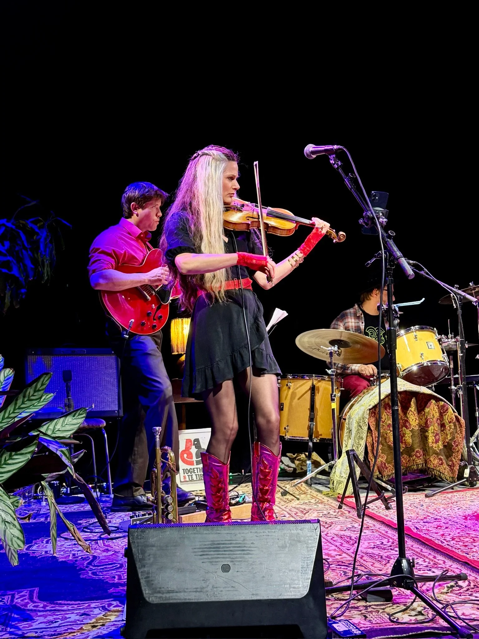 Three musicians performing on stage: a woman playing the violin and wearing red boots, a man playing electric guitar, and another person playing drums, with black background.