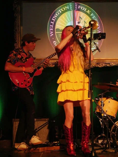 Female singer with long pink and yellow hair wearing a yellow ruffled dress and red boots, singing into a microphone on stage with a guitarist to her left playing an electric guitar.