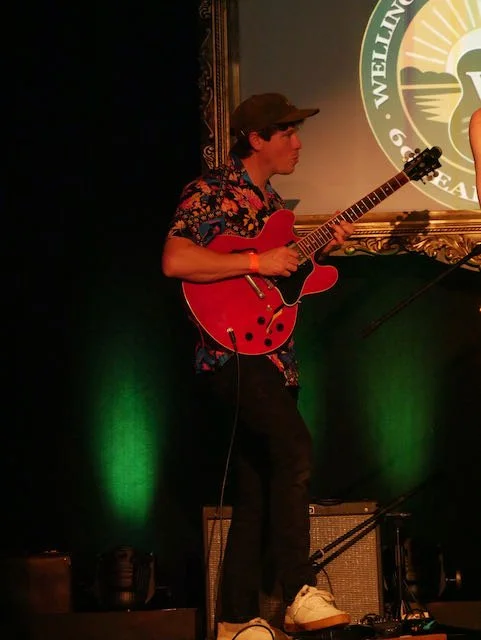 Young man in a floral shirt playing a red electric guitar on stage with green lighting and a logo in the background.