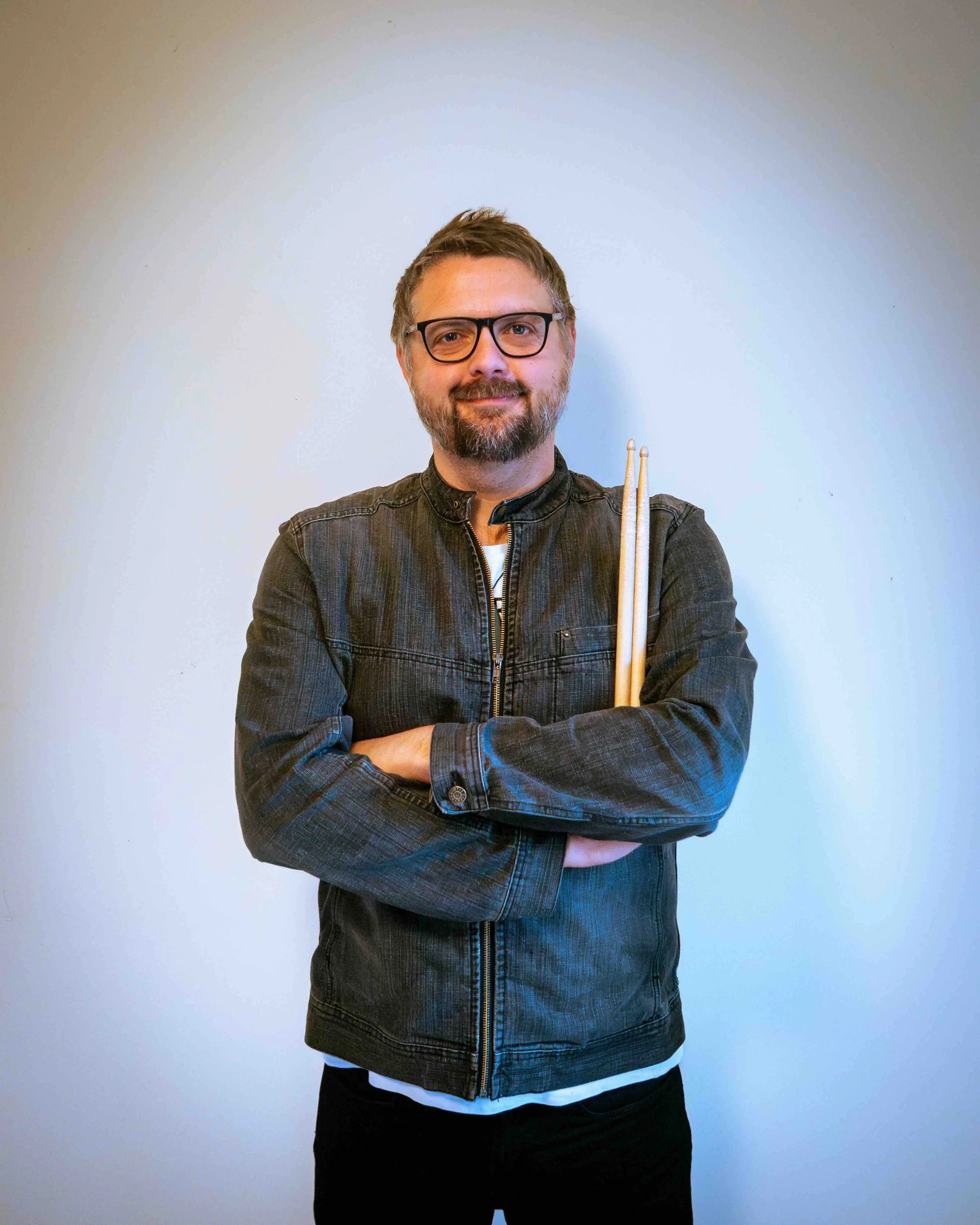 A man with glasses and a beard holding drumsticks cross-armed, wearing a black jacket against a plain light-colored wall.
