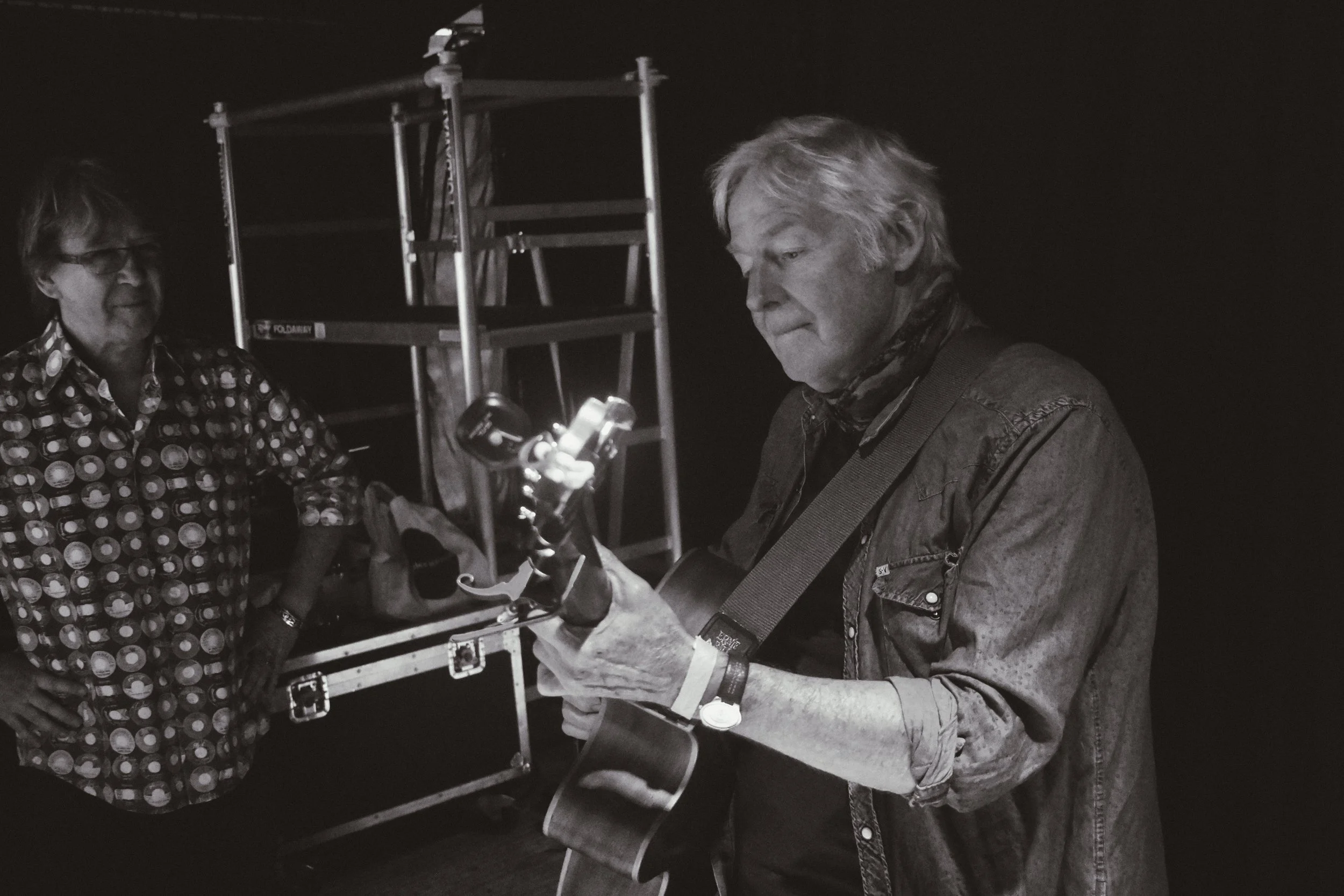 A man with gray hair wearing a denim jacket and scarf playing an acoustic guitar, with a woman in a patterned shirt standing nearby against a dark background.