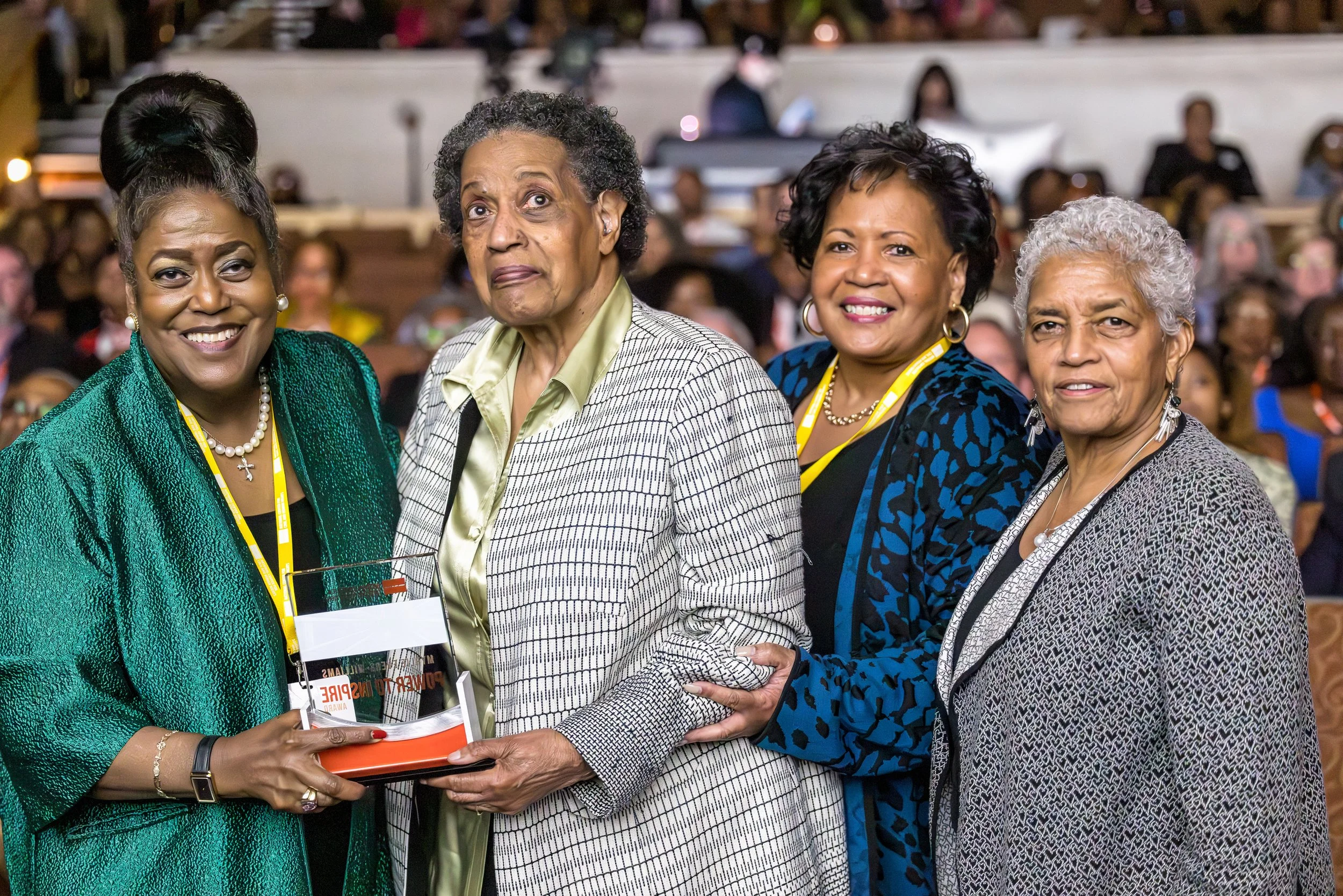 Four women of African descent standing together at an event, with one holding a plaque or award, and everyone smiling. There are many people seated in the background.