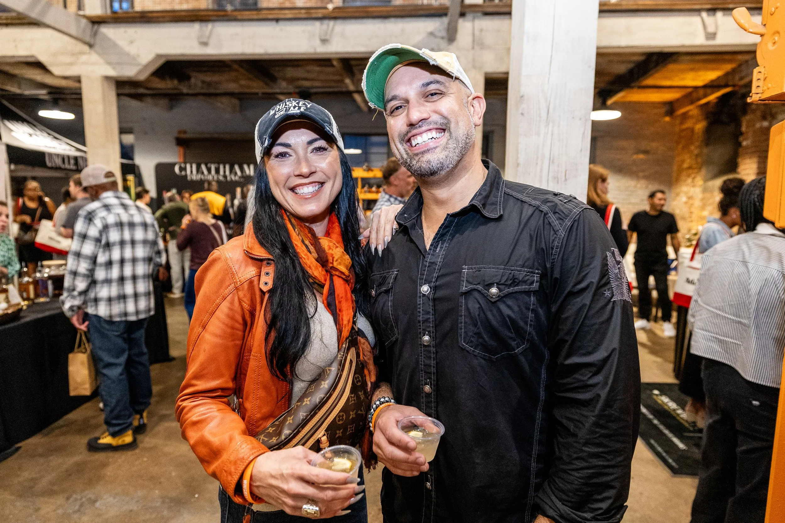 A smiling woman and man holding drinks at an indoor social event, with other people in the background.