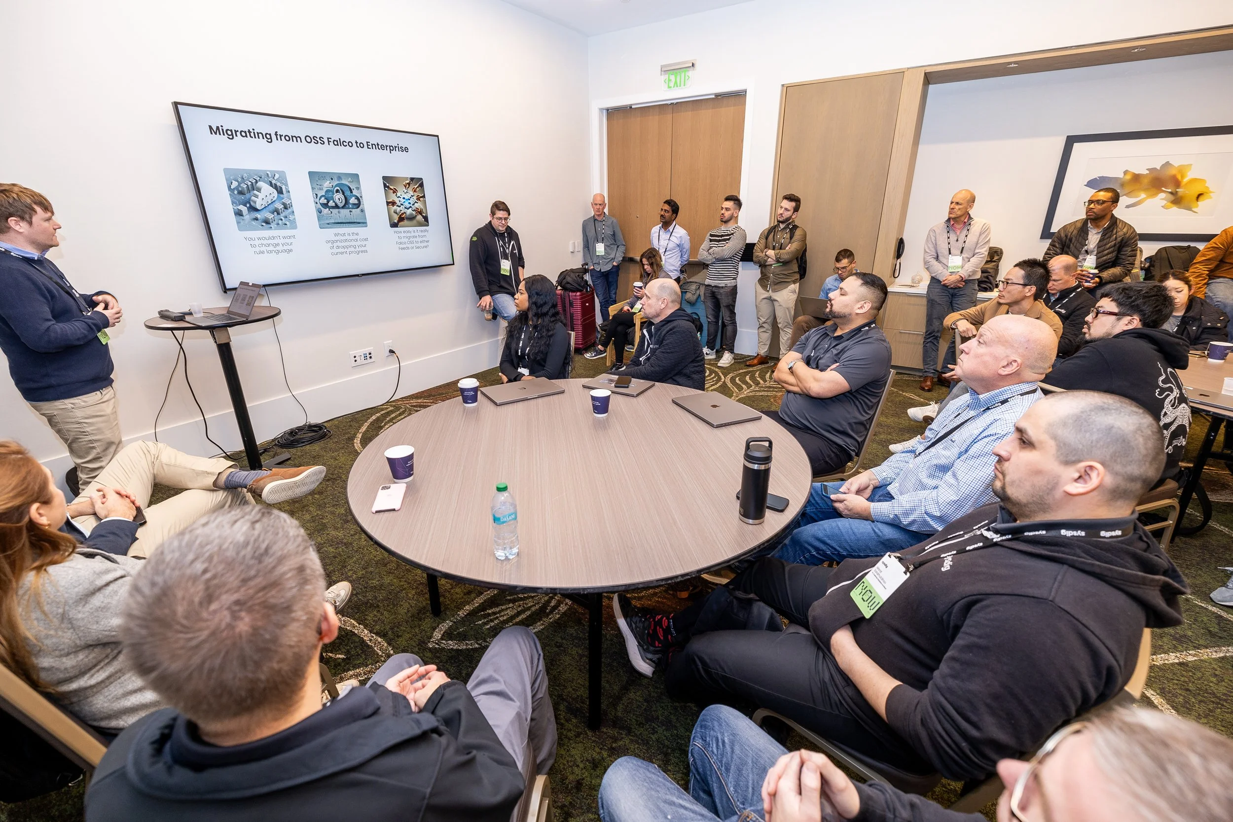 A group of people attending a presentation in a conference room, with a presenter standing near a large screen displaying a slide about migrating from OSS Falco to Enterprise.