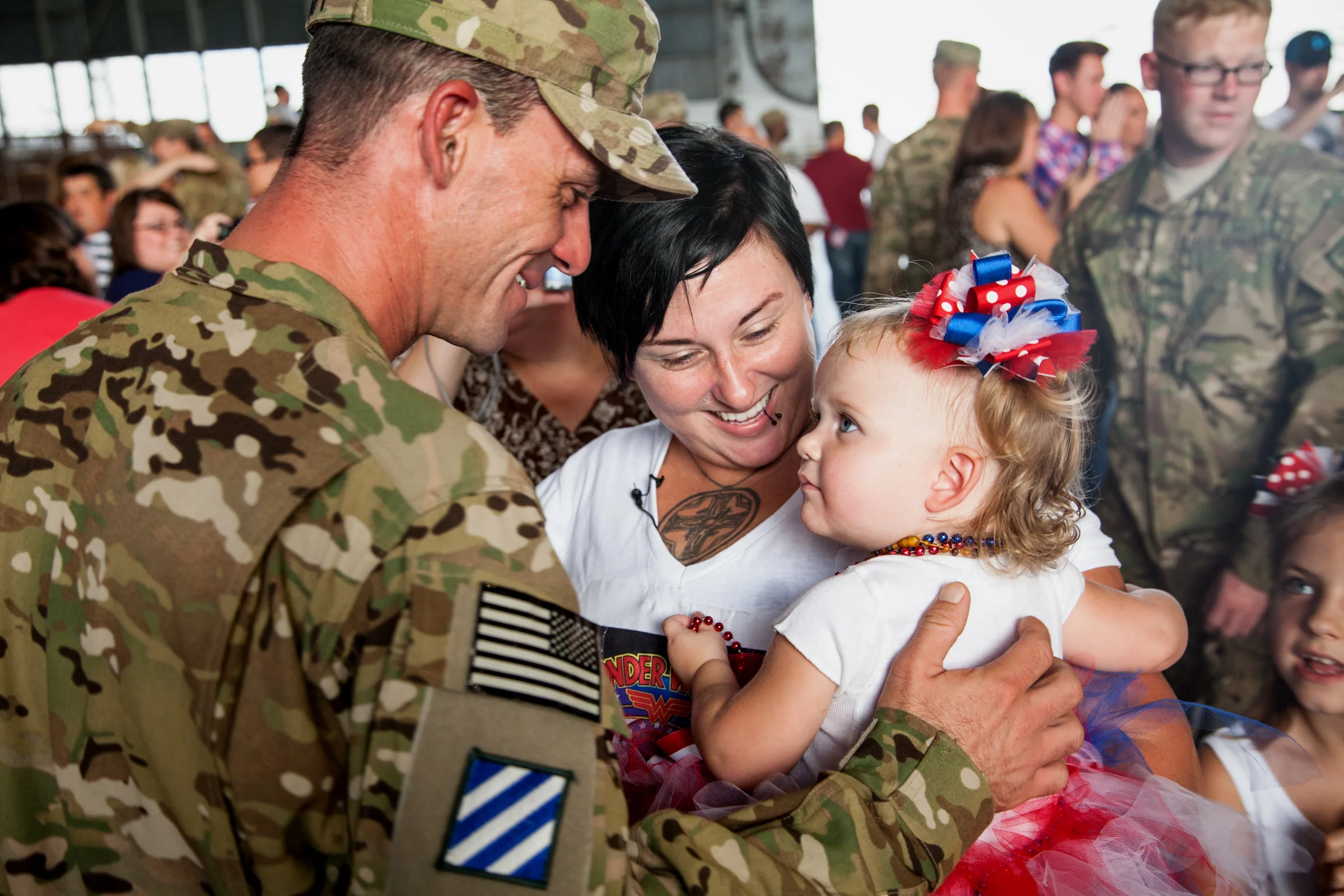 A soldier in camouflage uniform happily greets a young girl with a colorful bow in her hair at a gathering, surrounded by smiling people.