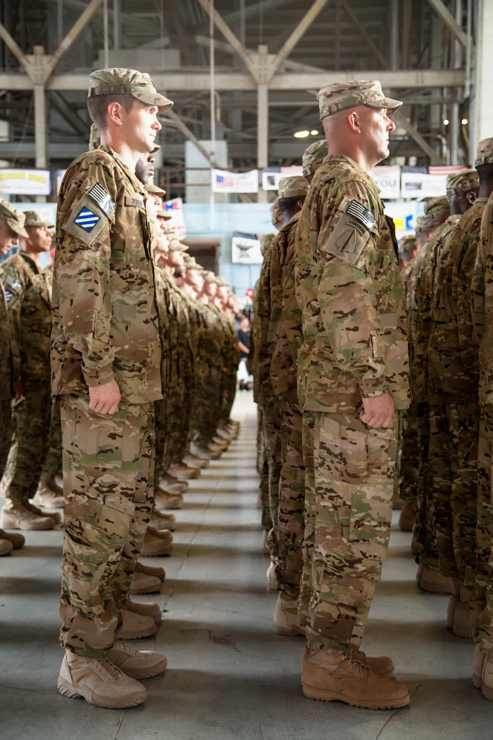 U.S. military soldiers standing in formation inside a hangar or warehouse, dressed in camouflage uniforms and tan boots.