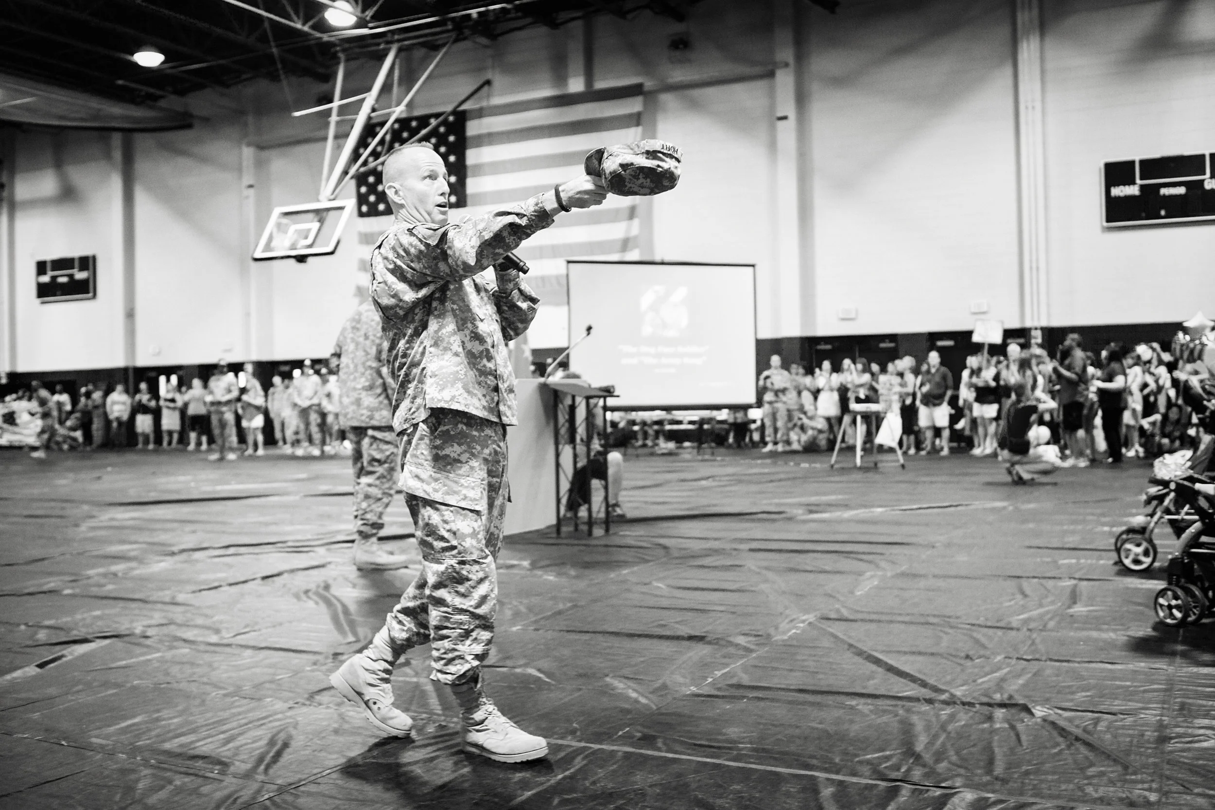A soldier in camouflage clothing is speaking into a microphone while holding a grenade shaped object at a military event in a gymnasium with a crowd of people and American flags in the background.