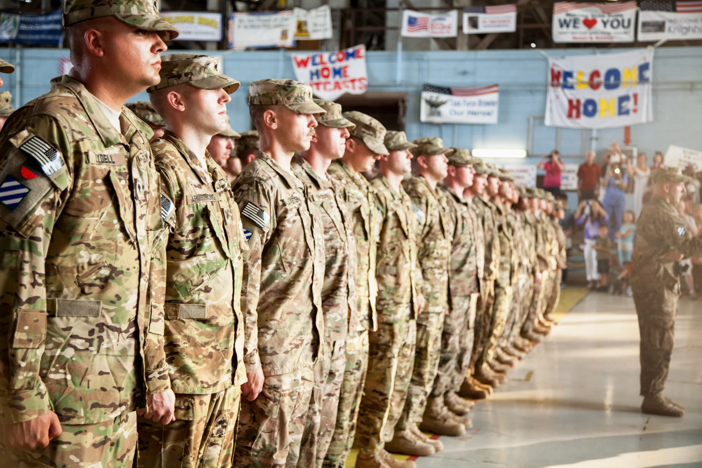 Military soldiers in camouflage uniforms standing in formation inside a building decorated with patriotic banners and signs that say "Welcome Home" and "We Love You."