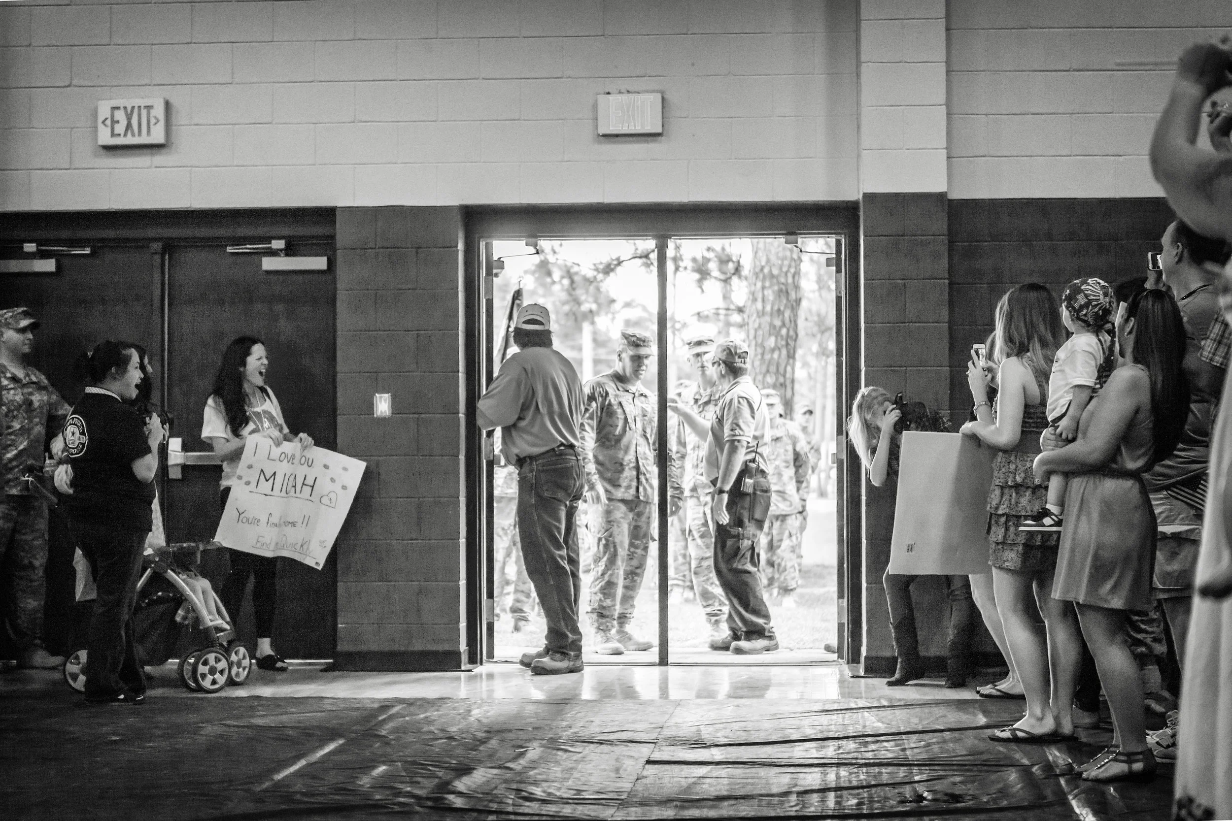 Military personnel and civilians inside a building, with some outside near the doorway. A woman in uniform holds a sign that says 'Love you MIAH. You're Awesome! Find Austin!'. Several people are taking photos or videos, and others are watching as a 