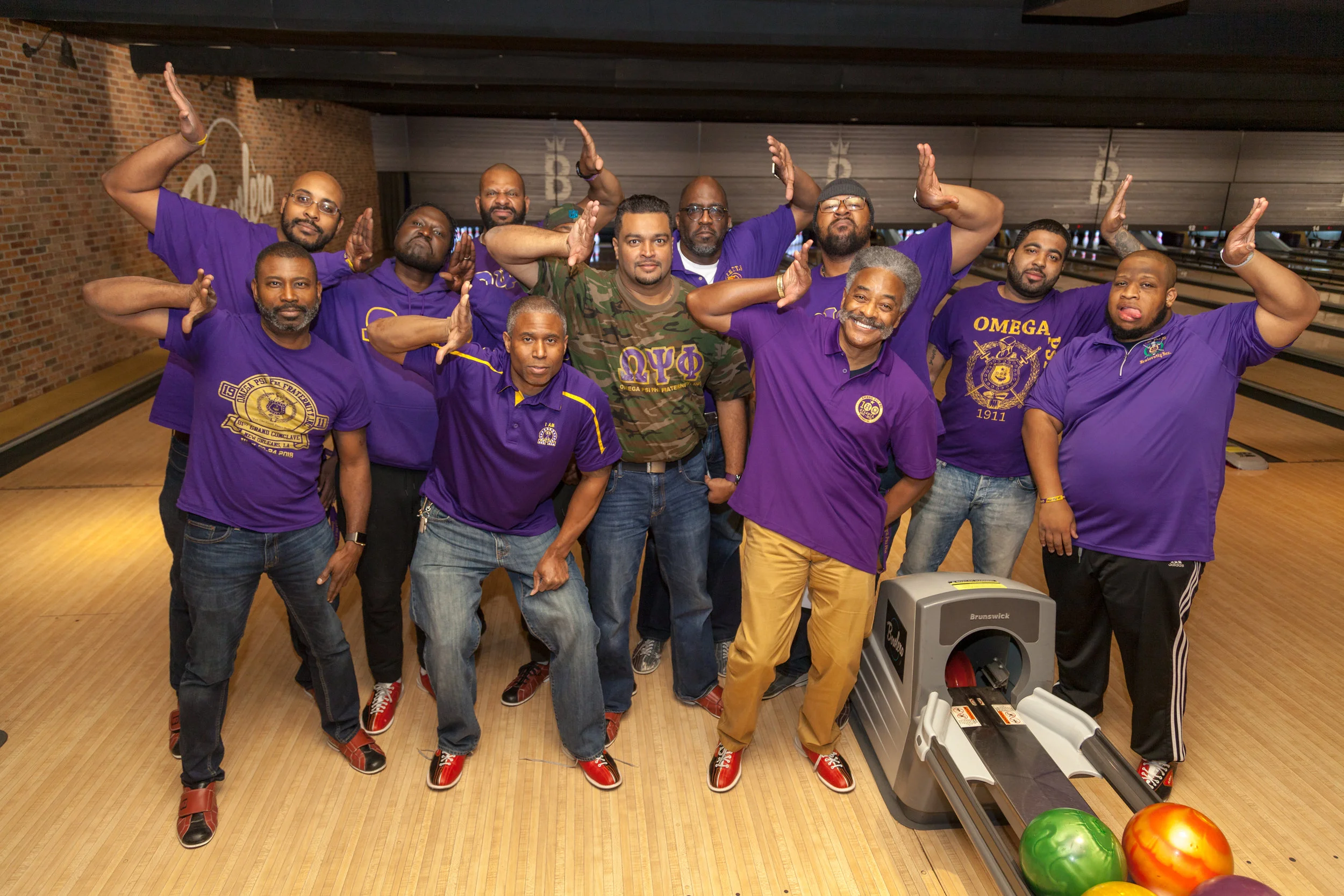 Group of men at a bowling alley, posing for a photo, some making peace signs. Many are wearing purple shirts with Greek letters and fraternity logos. Bowling balls and a ball return are visible in the foreground.