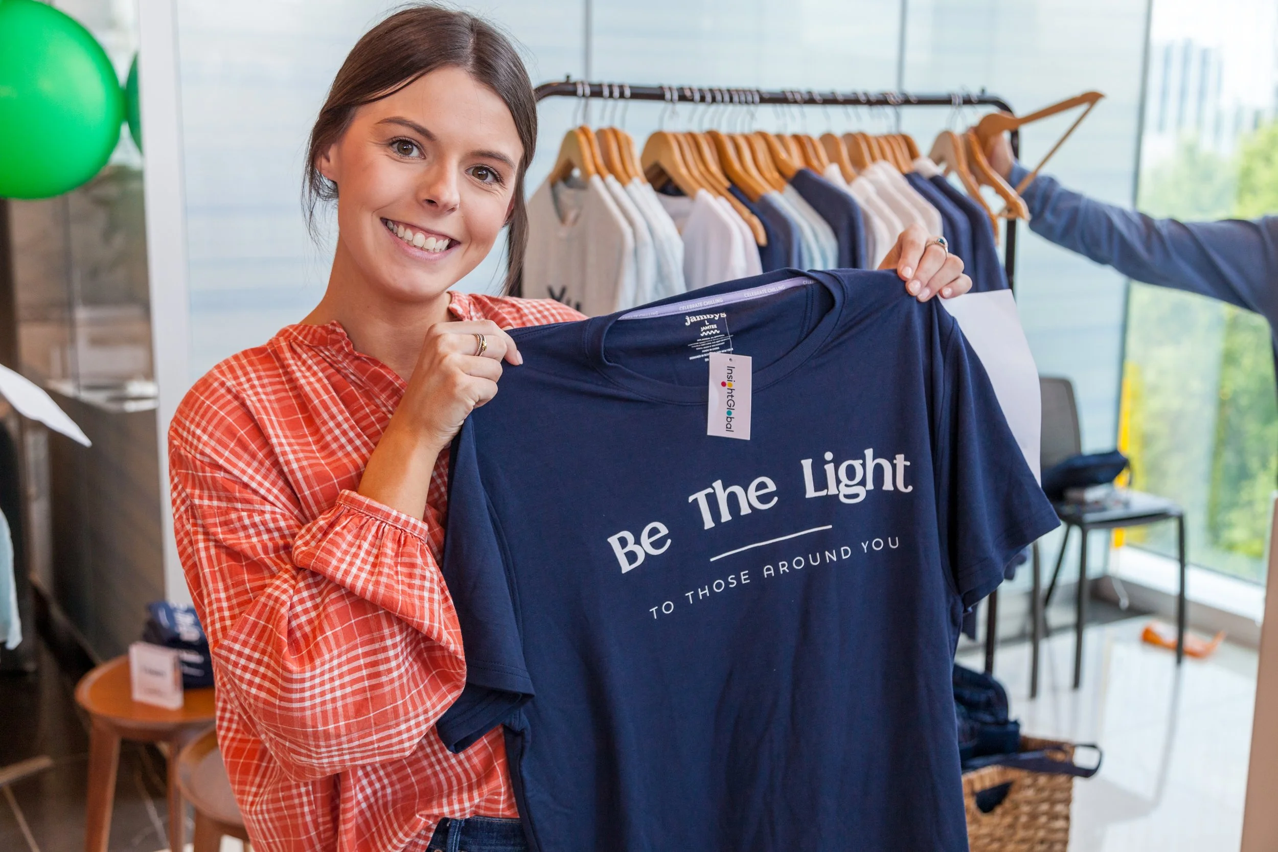 A woman with long brown hair smiling and holding up a navy blue T-shirt that says "Be The Light to Those Around You" inside a clothing store.