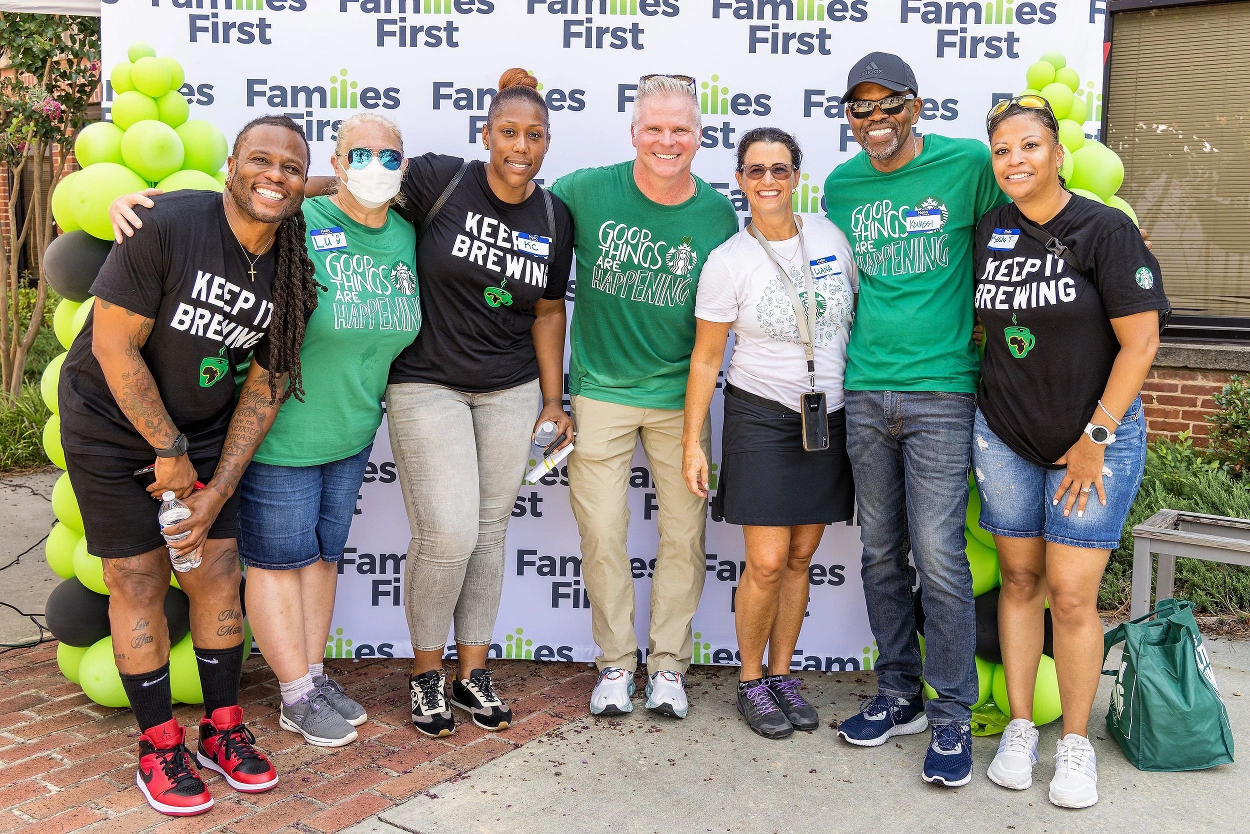 Group of seven diverse people standing in front of a backdrop with 'Families First' text, some wearing green shirts with 'Good things are happening' slogan, smiling, and holding water bottles.