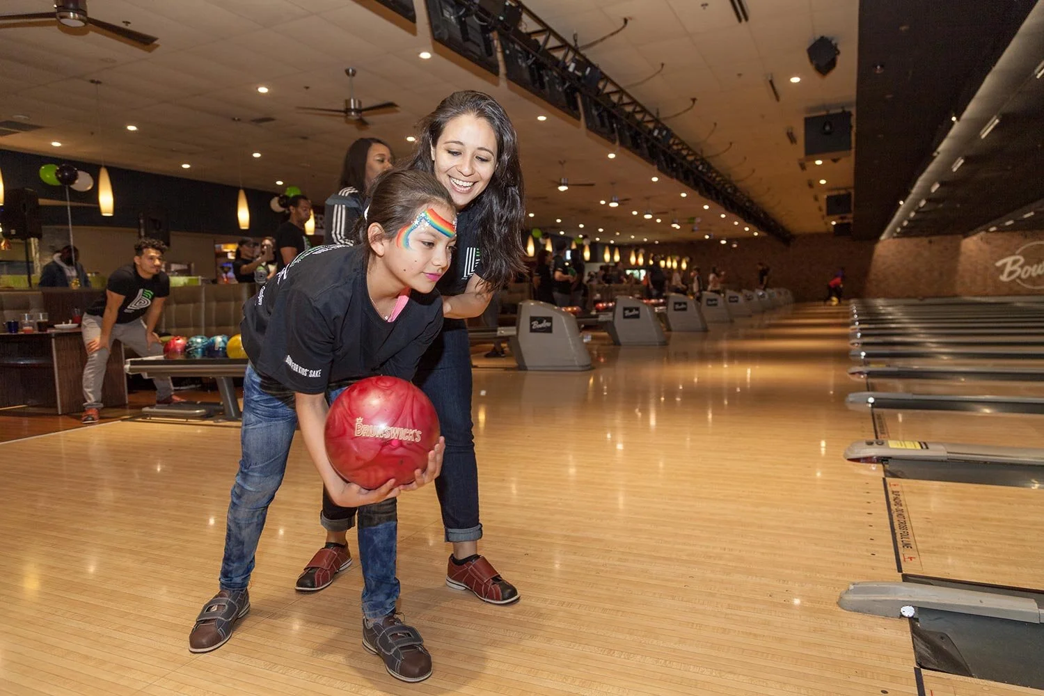Two women at a bowling alley, one helping the other with a red bowling ball, face paint on her face, surrounded by bowling lanes and other people.