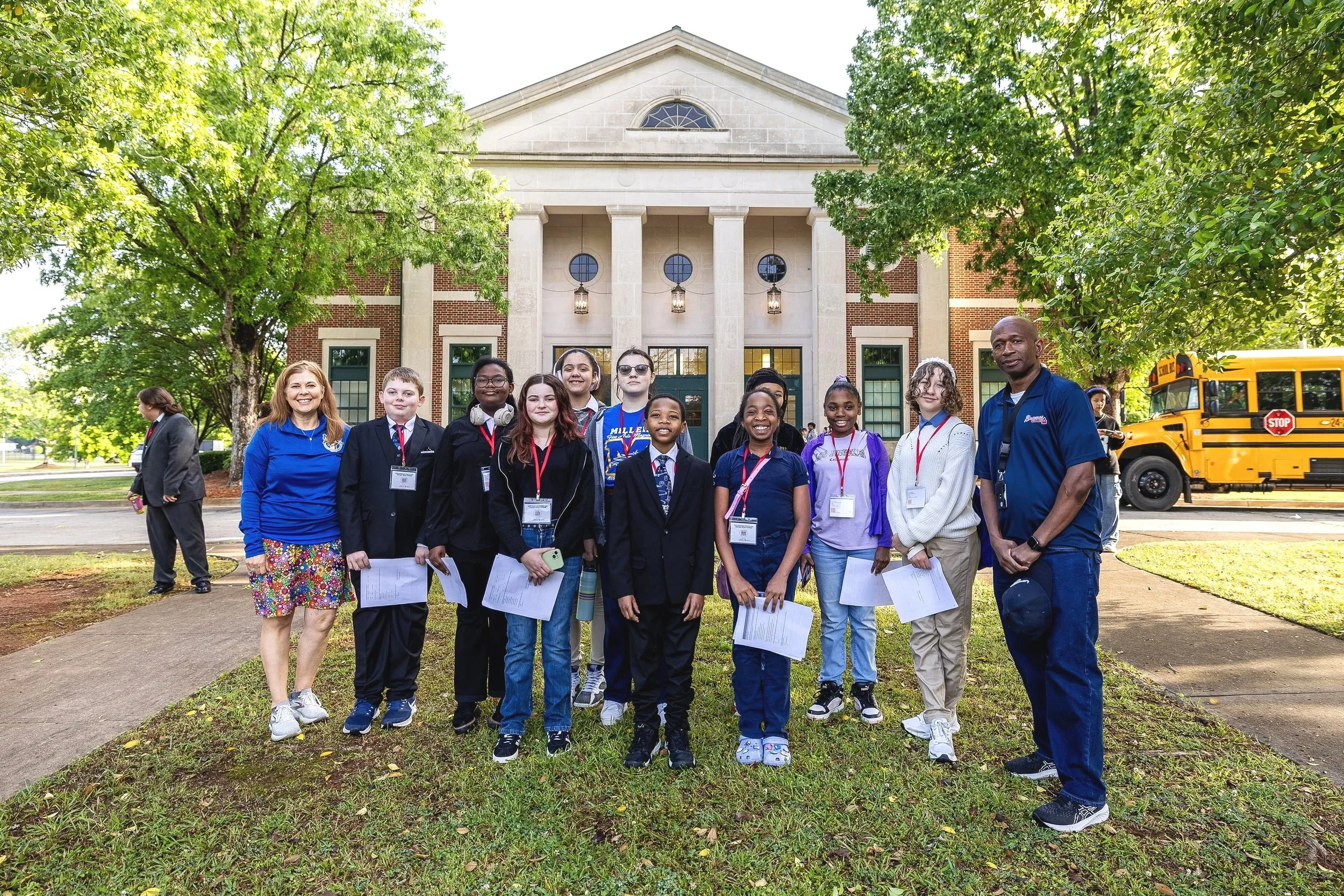 Group of children and two adults standing outside in front of a large building with trees and a school bus in the background. The children are smiling and holding papers, wearing ID badges, some dressed in formal clothes, others in casual attire.