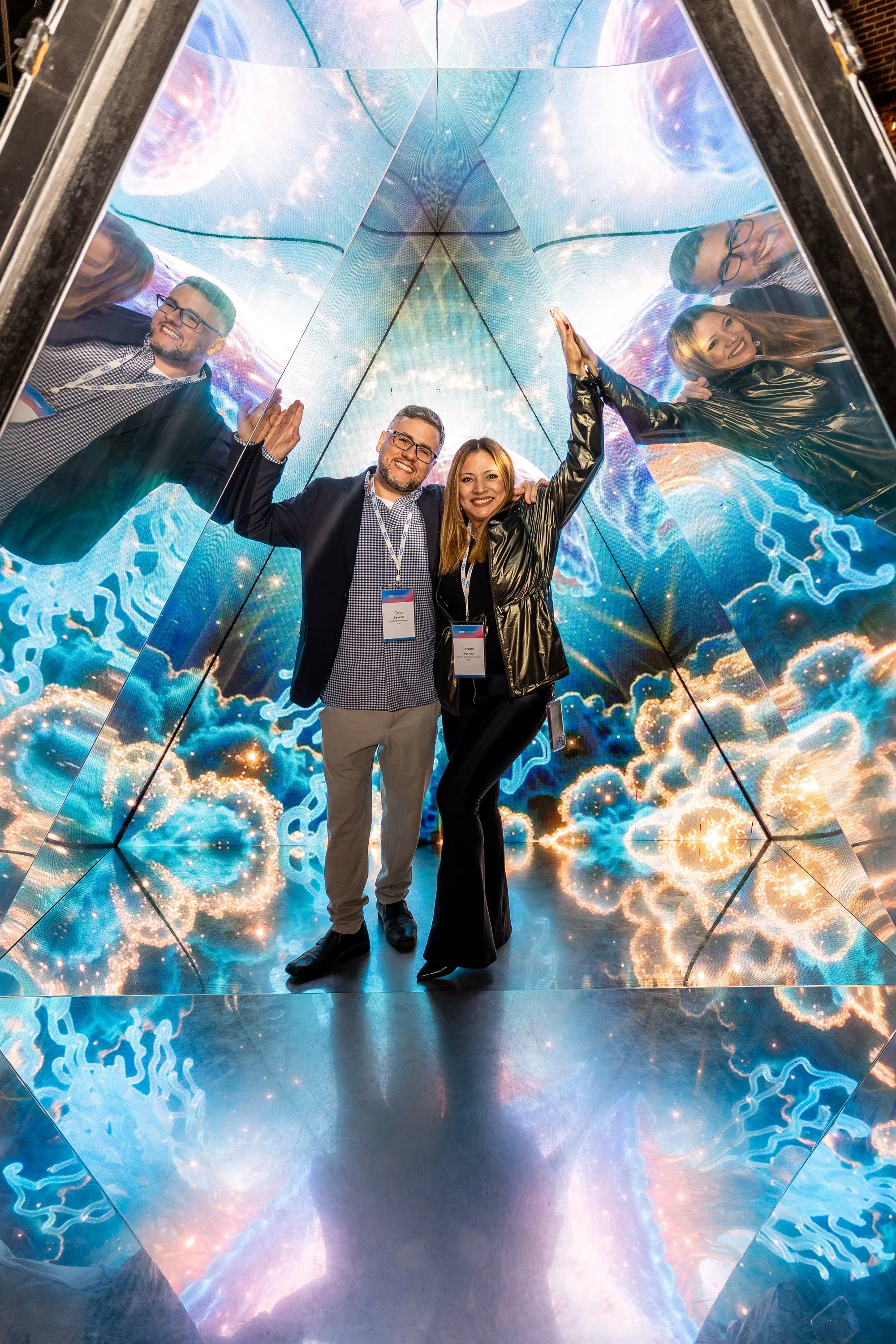 Two people, a man and a woman, are inside a reflective, mirrored cube with a colorful, abstract, cosmic pattern on the walls. They are smiling and high-fiving each other, with their reflections visible on the mirrored surfaces.