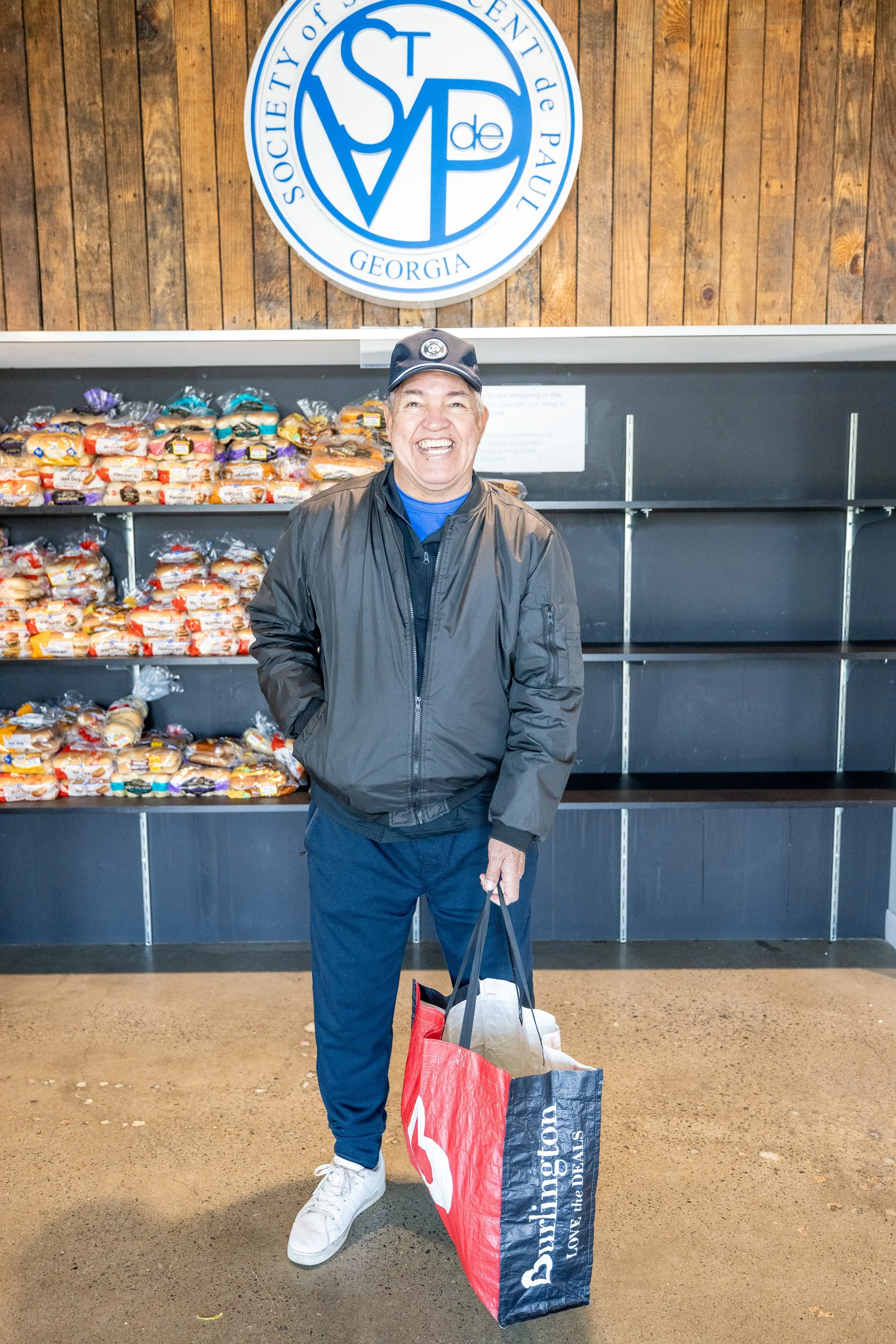 A smiling man wearing a black jacket, blue shirt, dark pants, white sneakers, and a baseball cap standing indoors in front of empty shelves and a government sign with blue lettering. He is holding a large red and black shopping bag. Behind him are sh