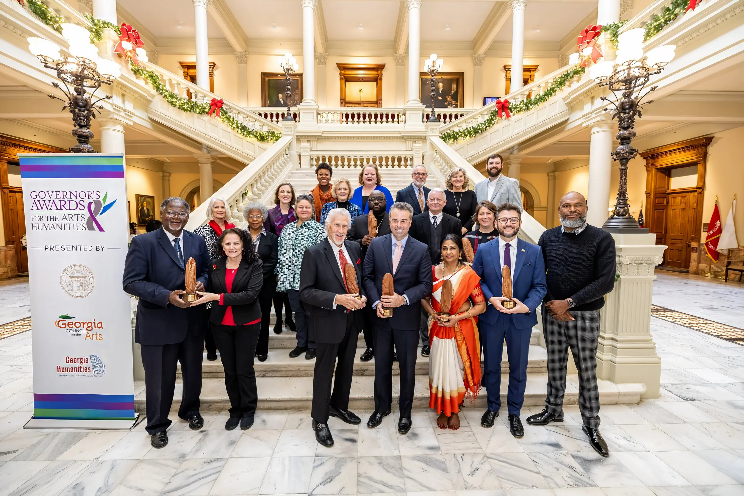 Group of people holding awards in front of a grand staircase at the Governor's Awards for the Arts & Humanities event, decorated with holiday garlands.