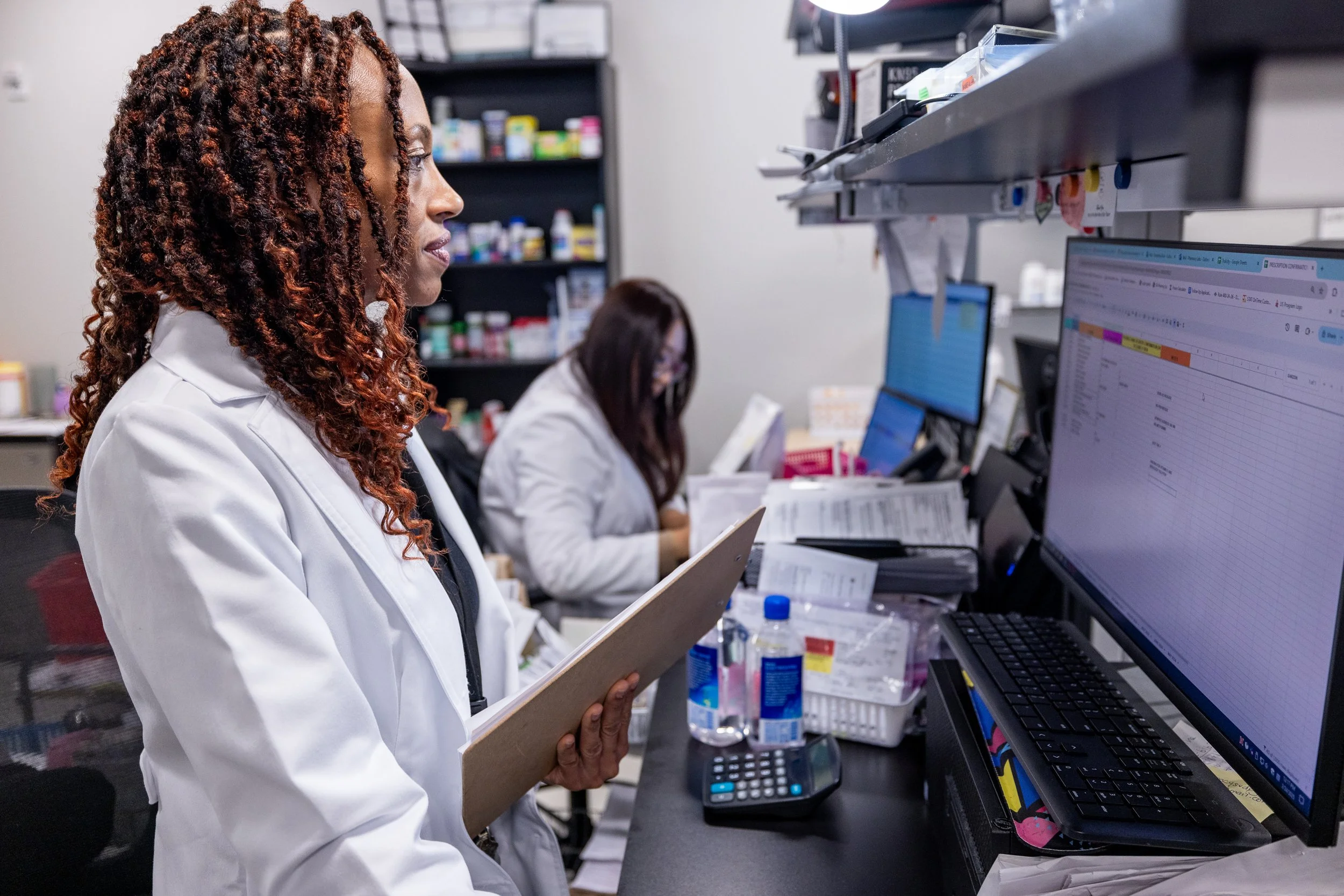 Two women wearing white lab coats working at a busy medical office or pharmacy, with one holding a clipboard and the other working on a computer, surrounded by medical supplies and paperwork.