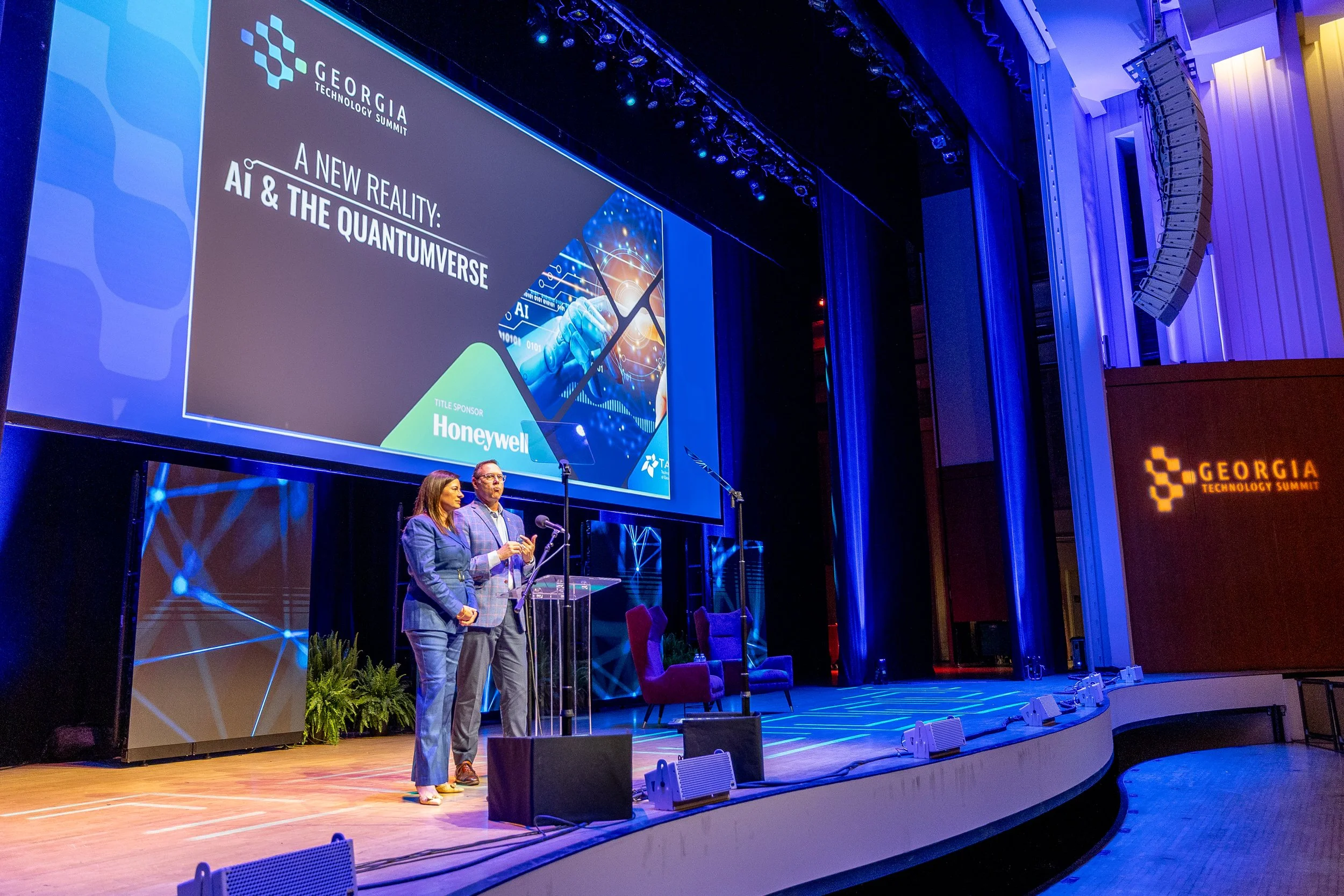 Two people speaking at a podium on a stage with a large screen behind them displaying the Georgia Technology Summit logo and a presentation titled 'A New Reality: AI & the Quantumverse.' The stage is decorated with plants and chairs, and the venue fe
