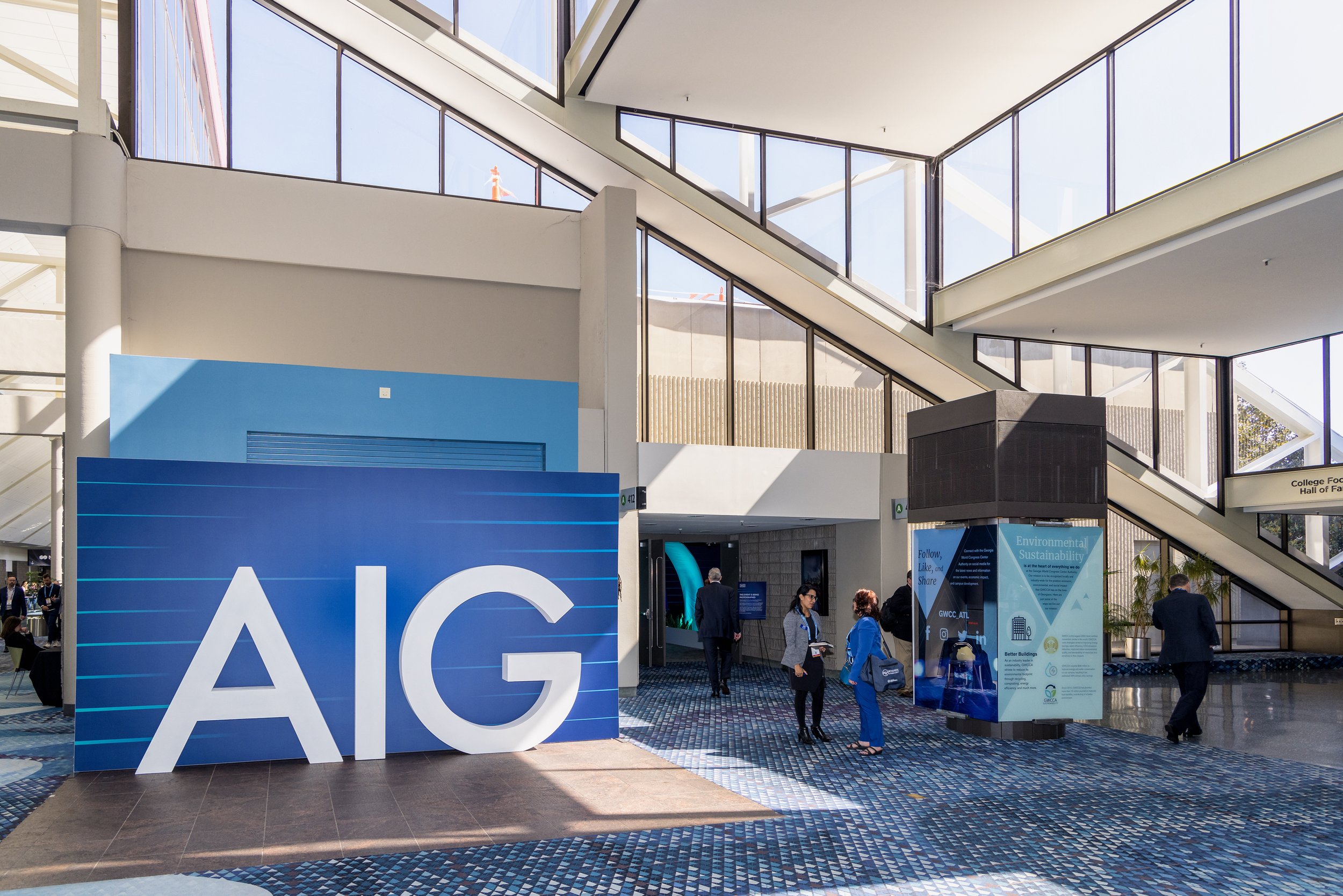 Indoor convention center with a large blue AIG sign, people walking and talking, and an informational display about environmental sustainability.