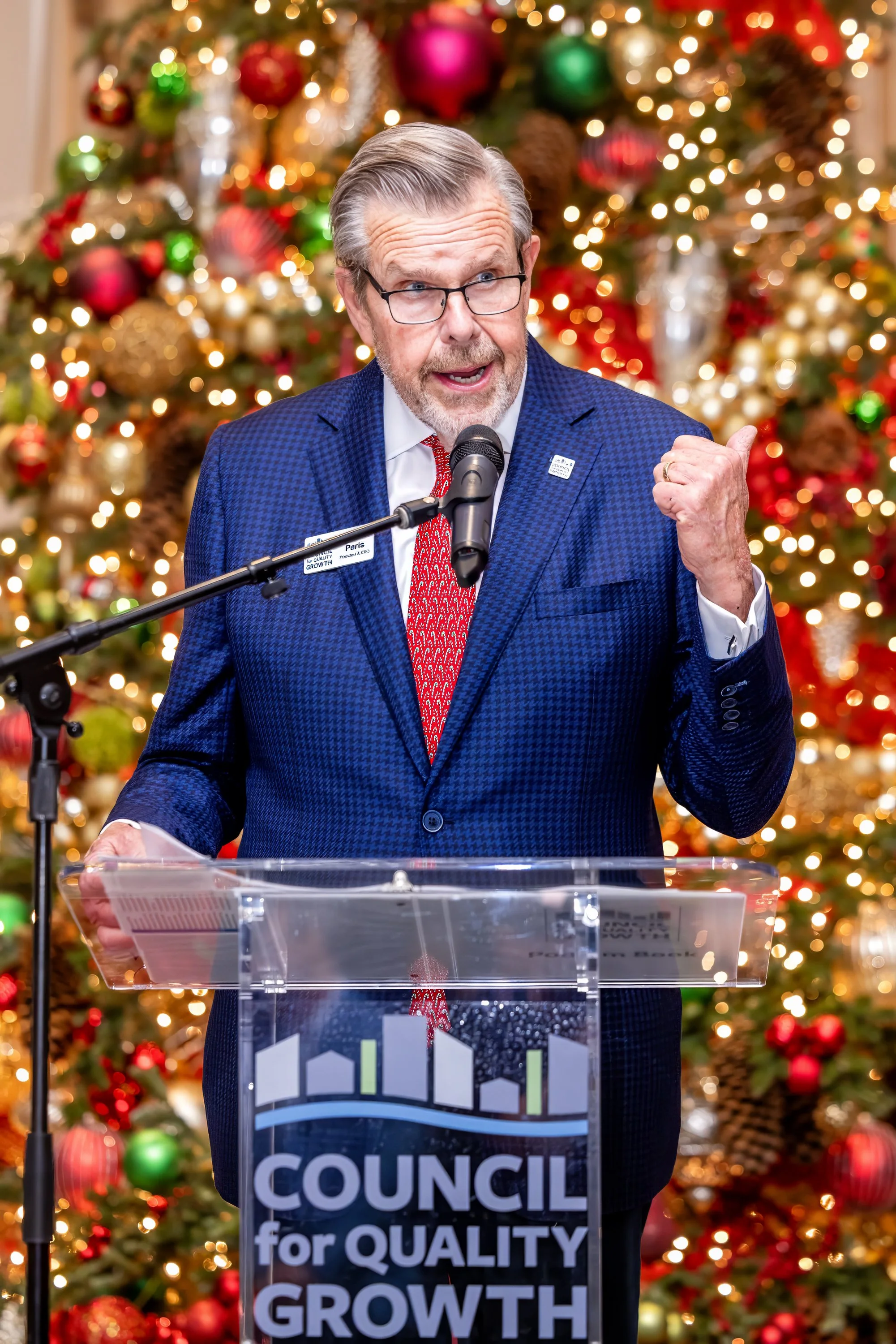 A man in a blue suit speaking at a podium with a Christmas tree decorated with ornaments and lights in the background.