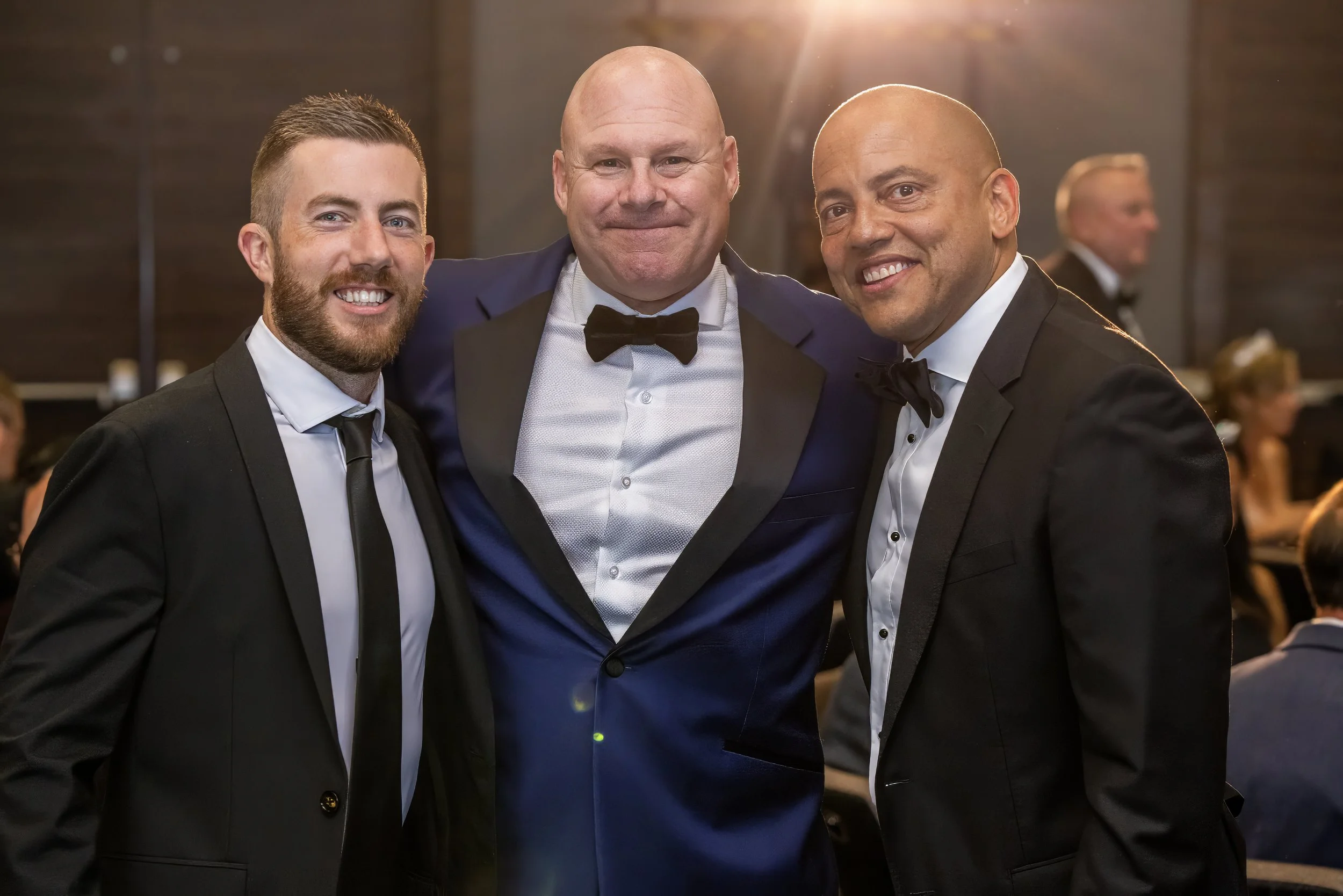 Three men dressed in tuxedos at formal event, smiling and posing together, with a blurred background of other guests.