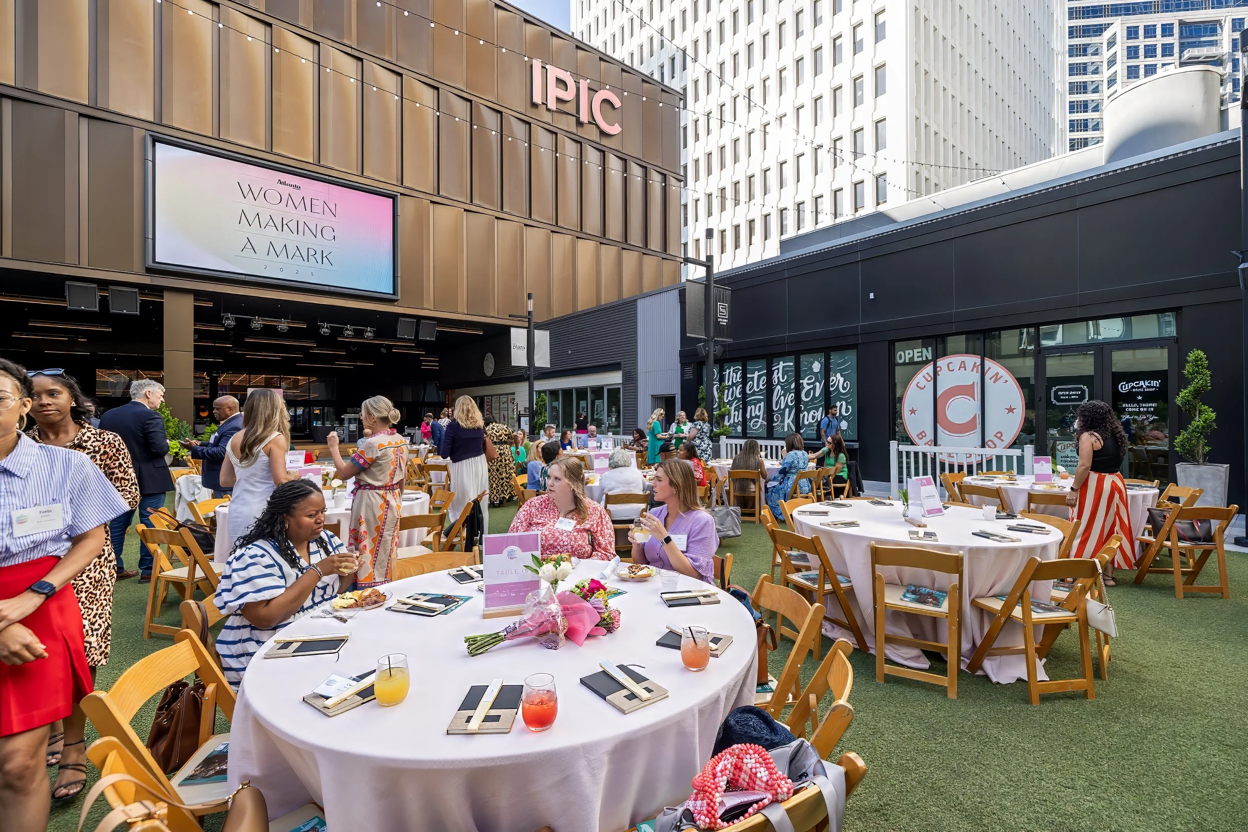 Outdoor event with round tables and chairs, people standing and sitting, some eating and drinking, a building with signs, and high-rise buildings in the background.