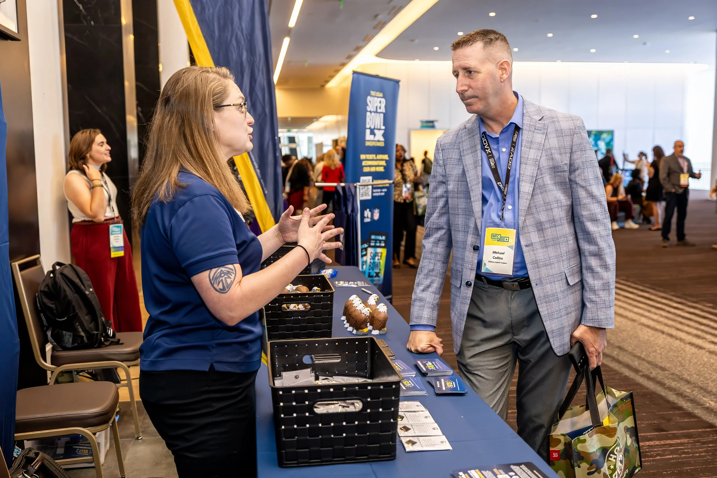 A woman in a blue polo shirt is talking to a man in a gray suit at a conference registration table. The woman has a tattoo on her left arm and glasses. The man is holding a camouflage tote bag. The background shows other attendees and a stage in a la