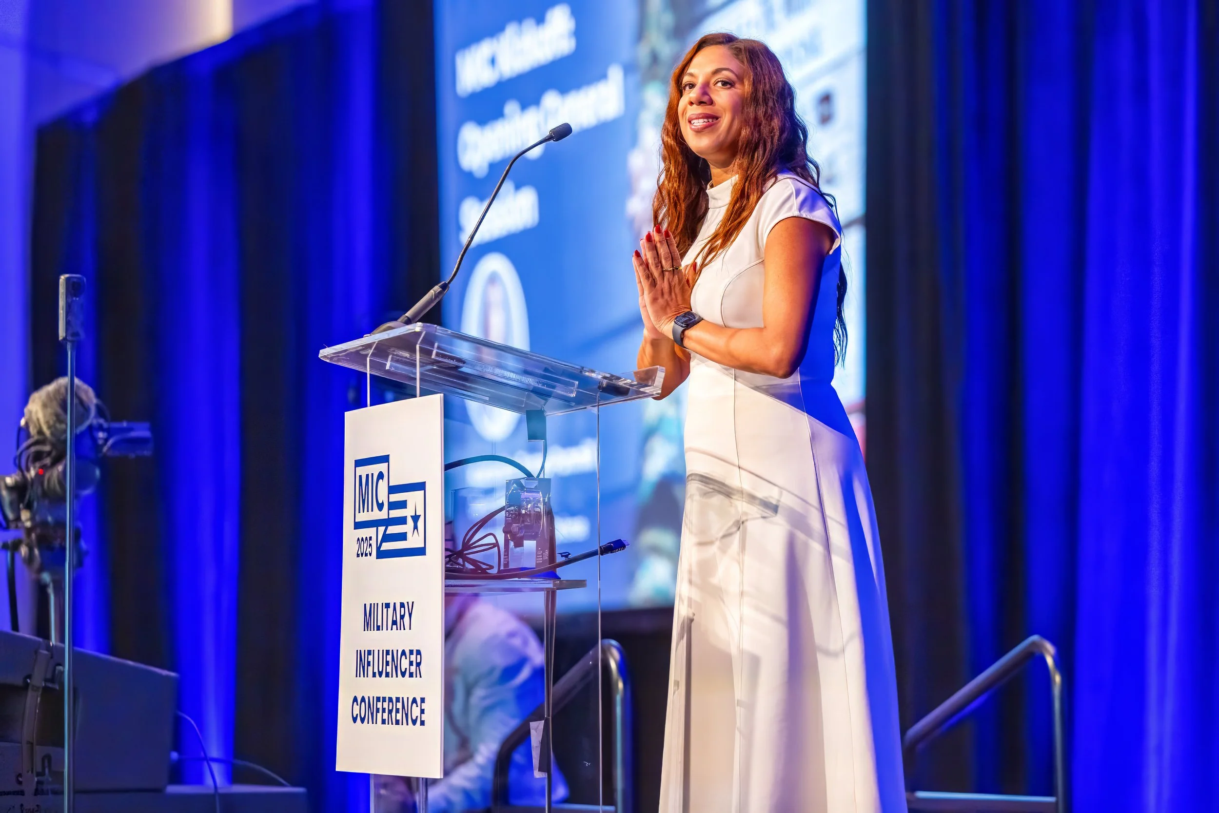 A woman in a white dress standing at a podium at the Military Influencer Conference 2025, with her hands pressed together and smiling, in front of a large blue and purple curtain backdrop.