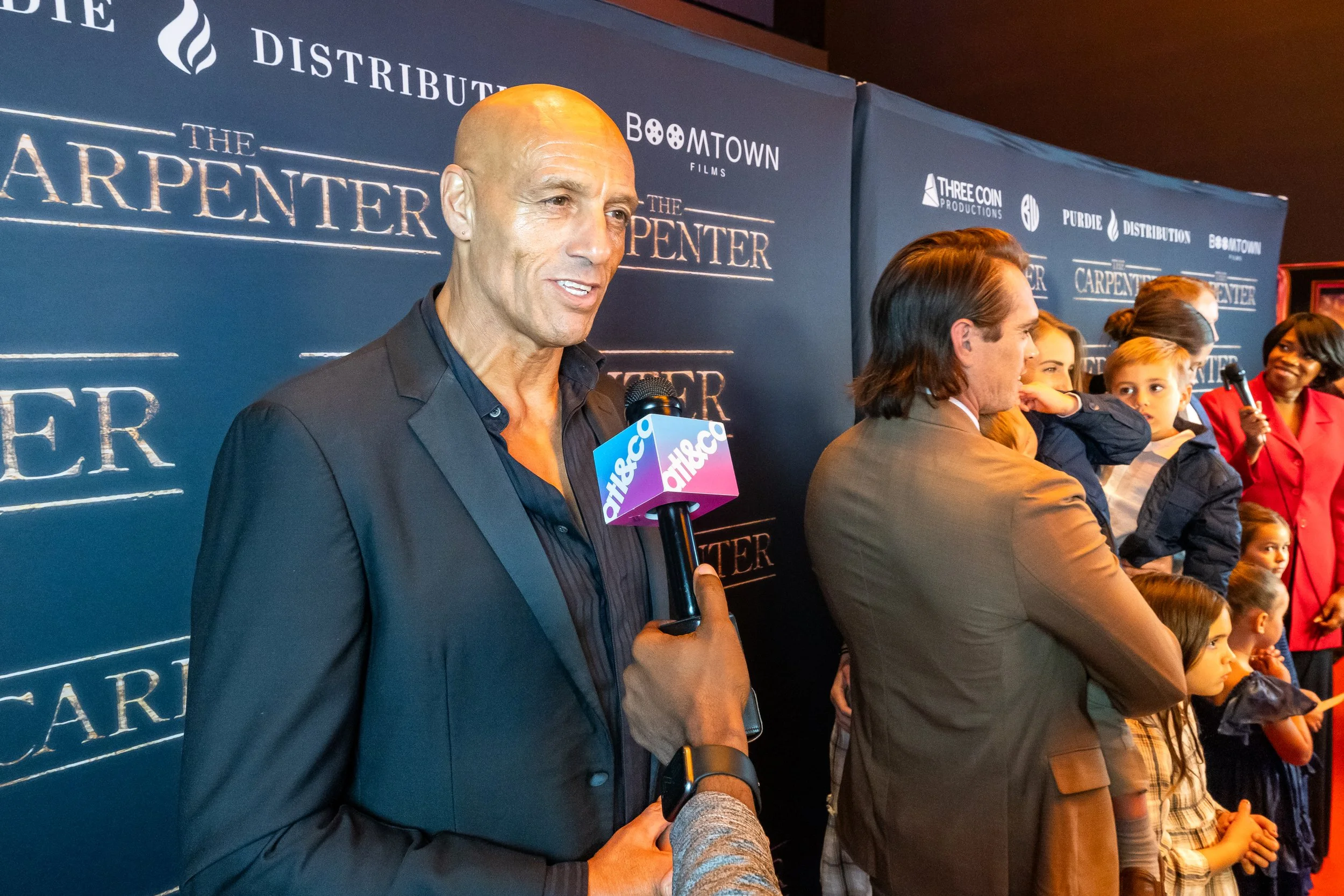 A man being interviewed at a red carpet event for the movie 'The Carpenter', with a microphone that has the logo 'alico'. Behind him is a group of people, including children and women, standing in front of a dark blue backdrop with the movie title an