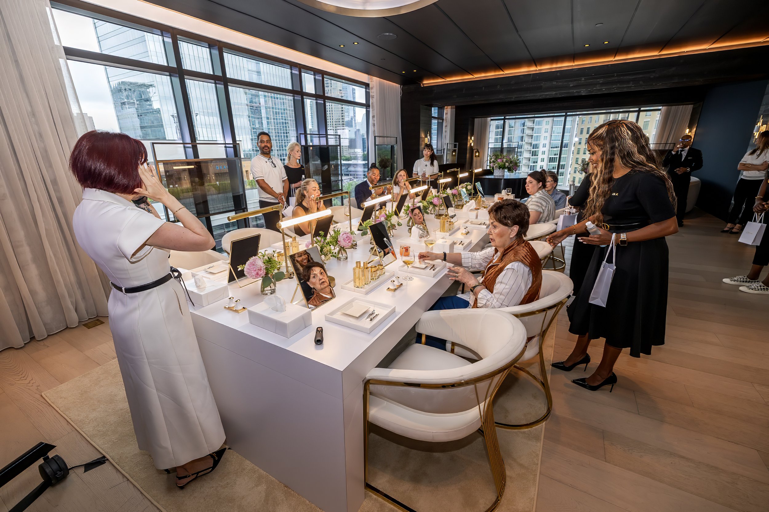 A group of women with beauty or fashion products in a luxurious, well-lit room with large windows showing city buildings; some women are seated at a table, while others stand around, engaging in a social or beauty event.