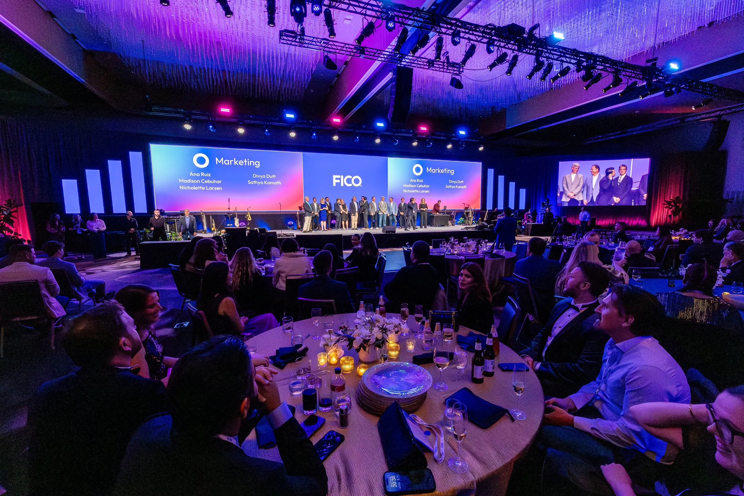 A large conference room filled with seated attendees watching a presentation on stage. The stage has several large screens displaying the word 'Marketing' and the names Ann Ruiz, Madison Cebuhar, Nicholettte Larsen, Divya Dutt, and Sathya Kamath, alo