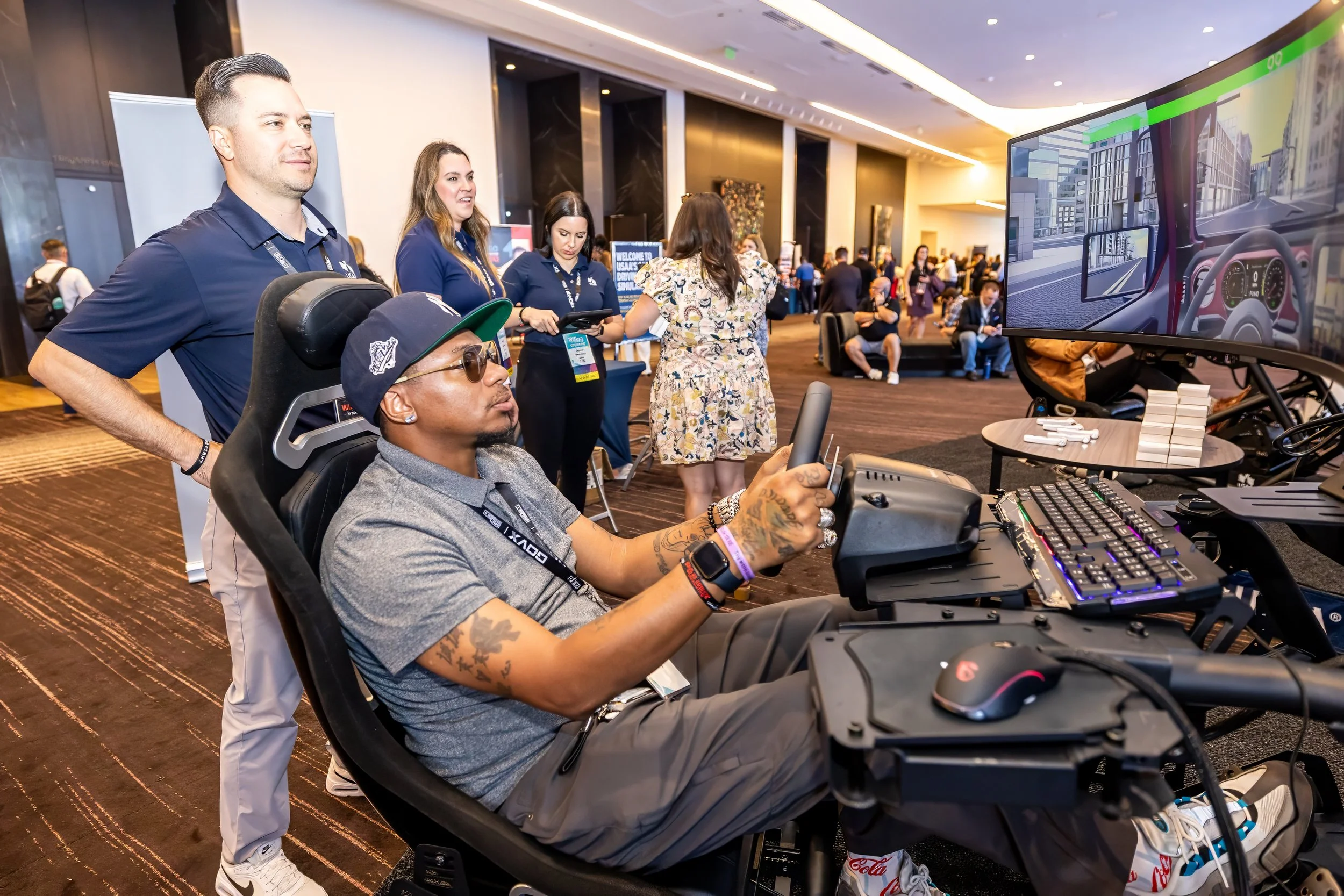 Person in racing simulator at a tech convention, surrounded by attendees and display booths.