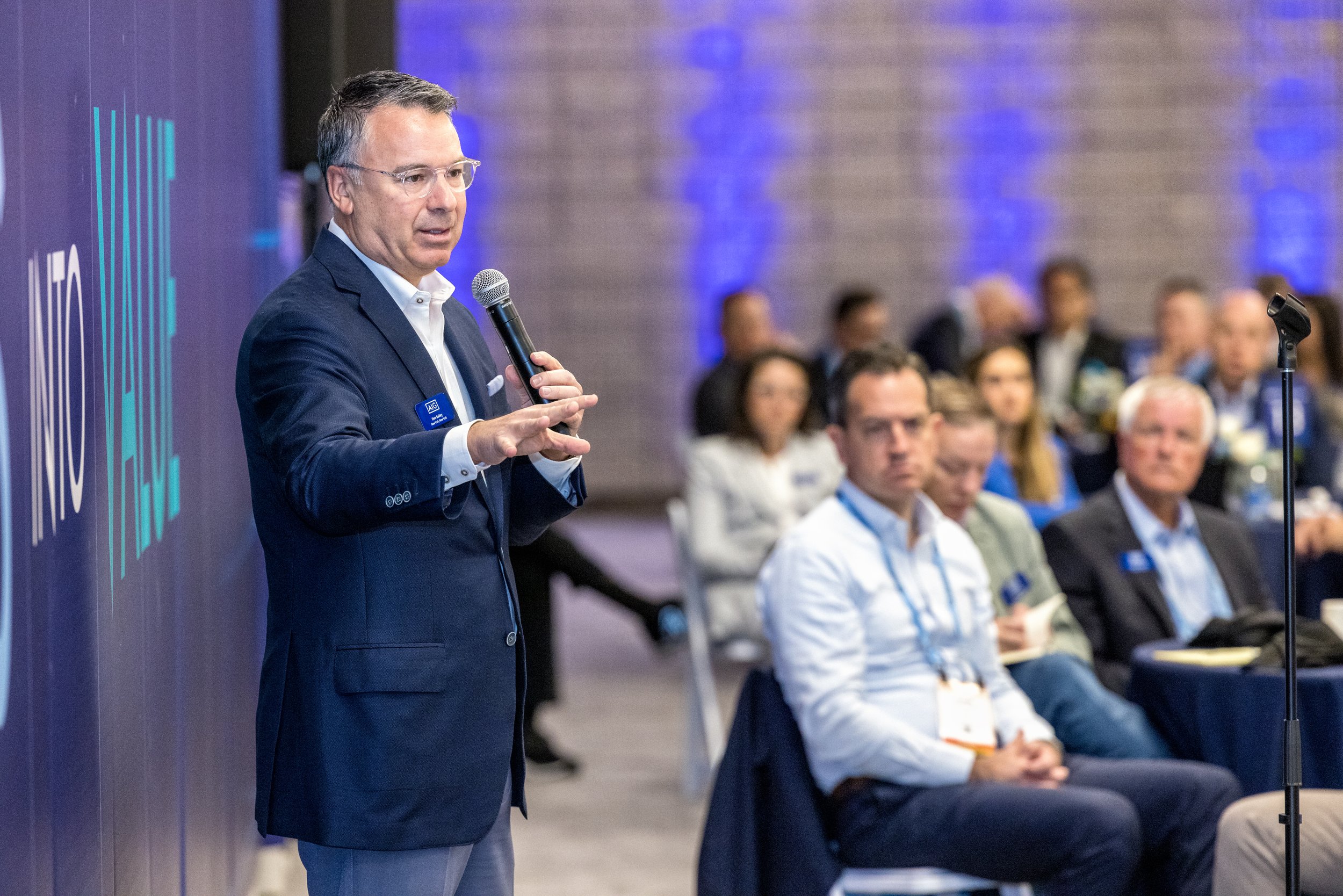A man in a dark blue suit and glasses is speaking into a microphone during a conference. He is standing in front of a blue backdrop with white text. The audience, consisting of men and women, is seated and listening attentively.