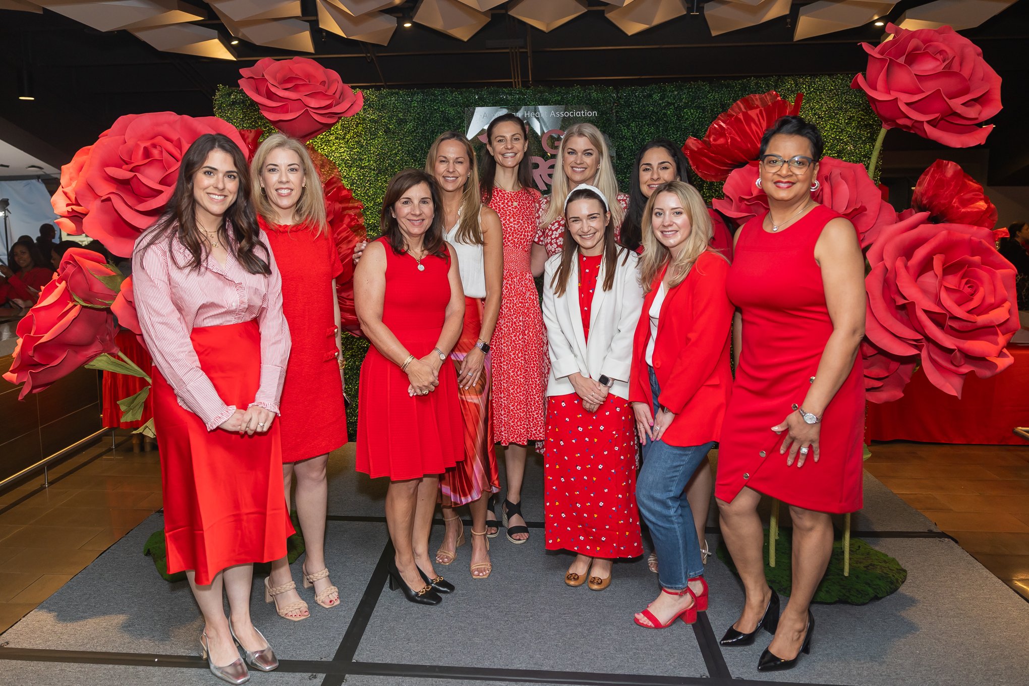 Group of eleven women standing together in front of a green backdrop with large red roses at an American Heart Association event, all smiling and dressed in red and white outfits.