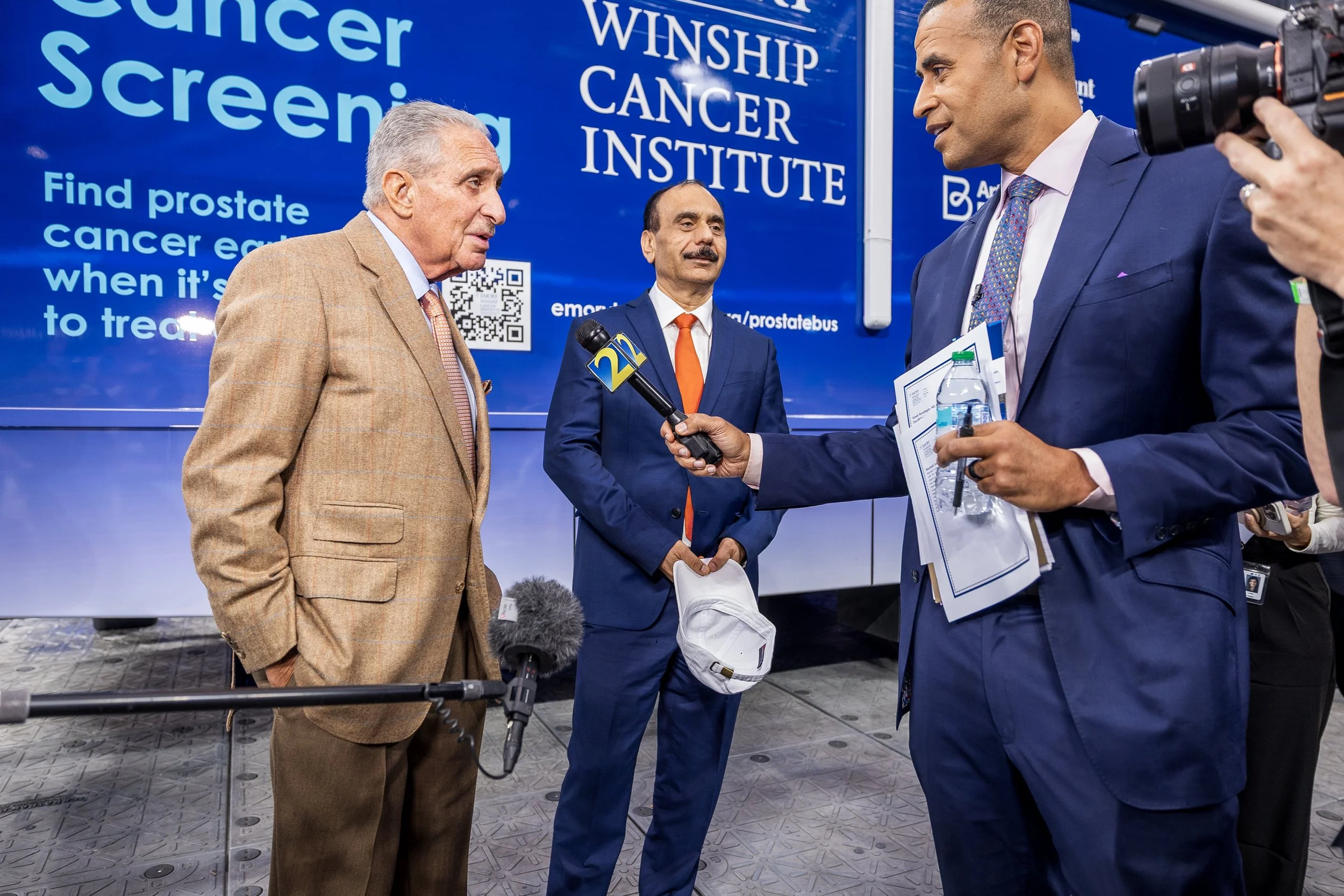 Three men in suits being interviewed in front of a large blue screen with the text 'Winship Cancer Institute' and a QR code, at an event related to prostate cancer awareness.