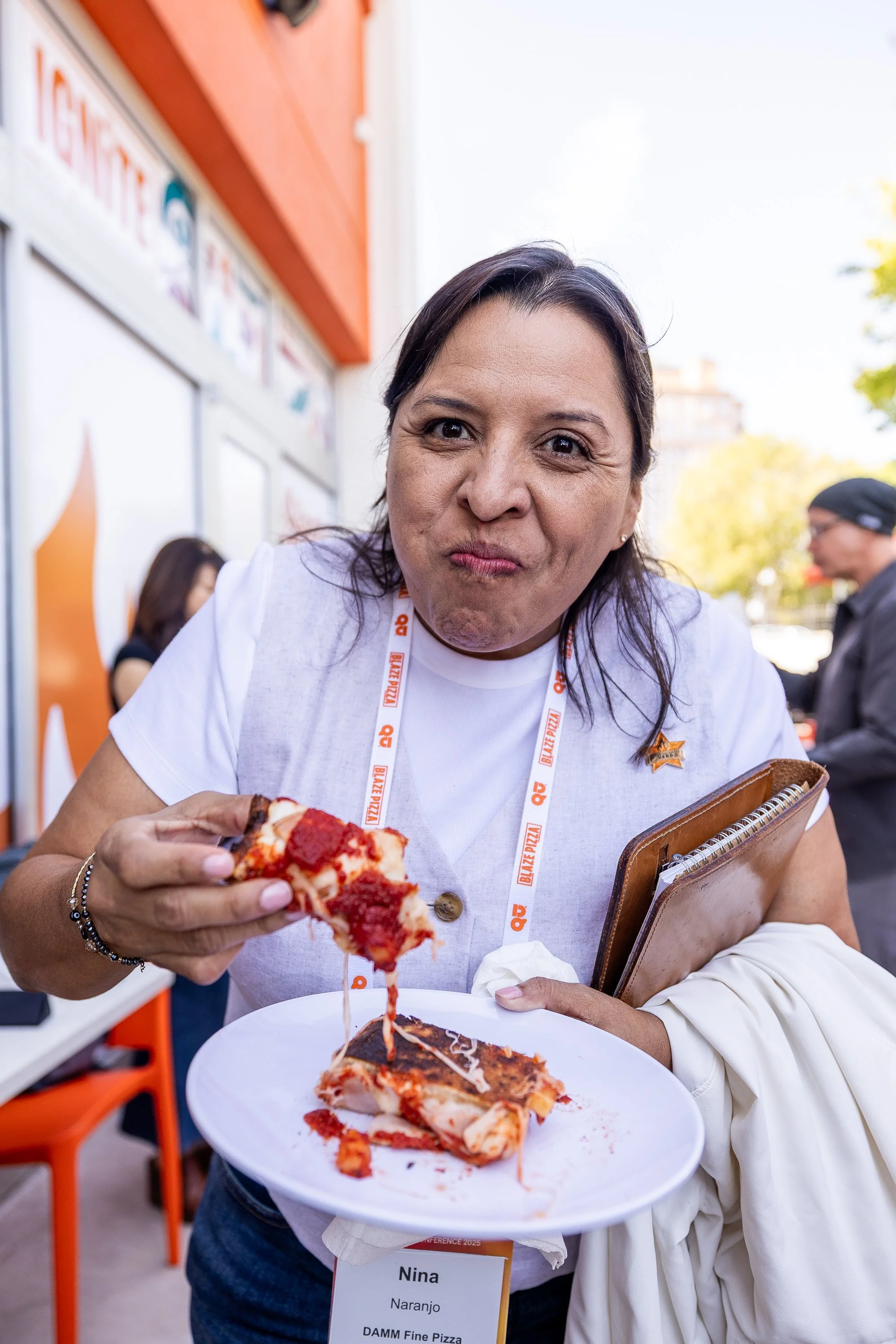 Woman enjoying a slice of pizza at an outdoor event, holding a plate with pizza and a notebook, wearing a white shirt with a name tag that says Nina