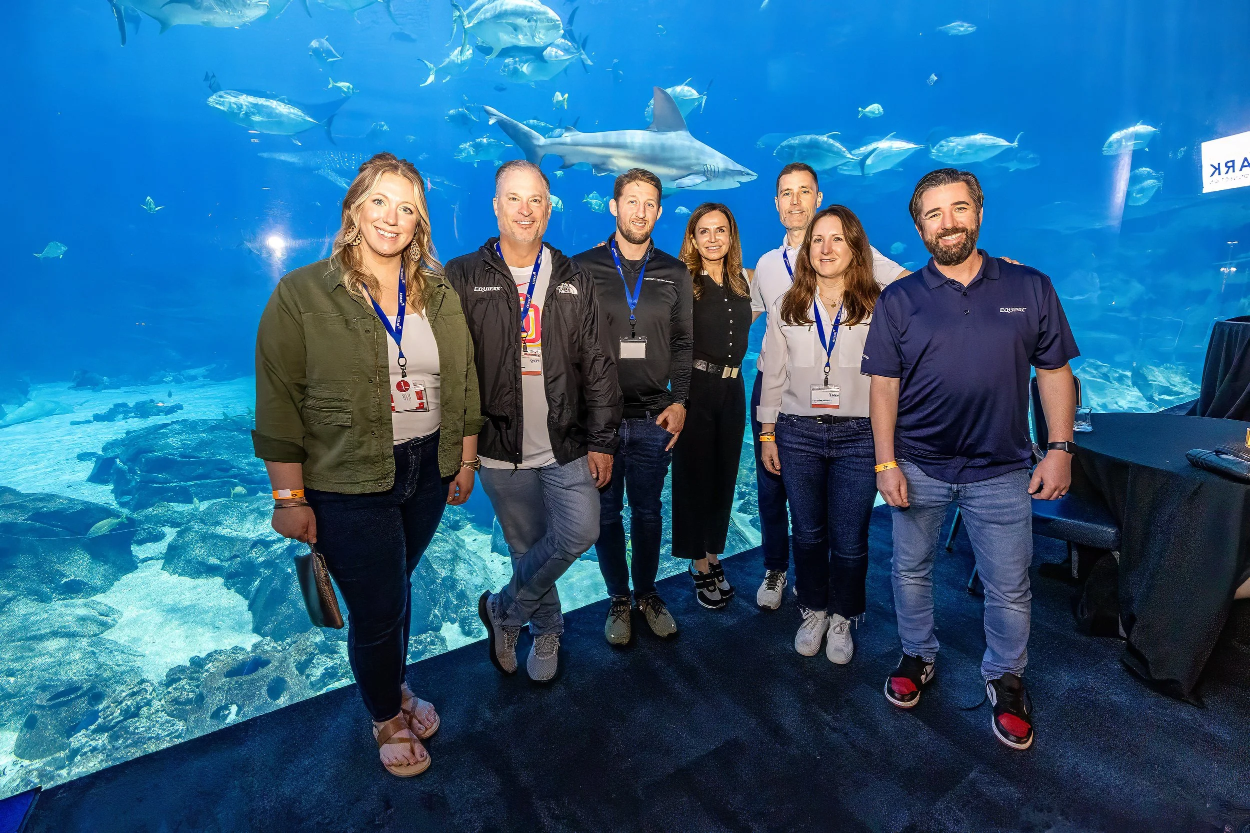 Group of seven people standing in front of a large aquarium tank with various fish and a shark swimming in the background.
