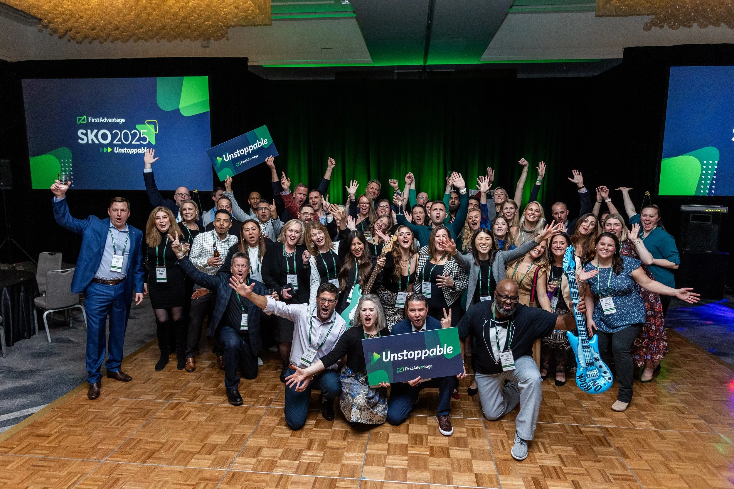 Group of people at a conference celebrating, holding signs that say "Unstoppable" and a guitar-shaped inflatable, with a large screen in the background displaying "First Advantage SKO 2025 Unstoppable."