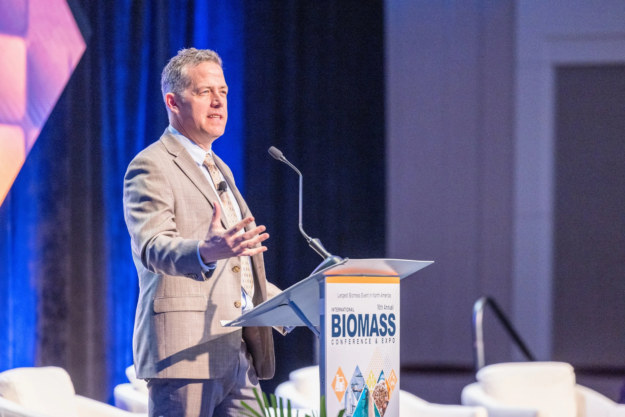 A man in a beige suit and tie speaking at a lectern during a conference, with a large blue and orange banner behind him that reads 'International Biomass Conference & Expo'.