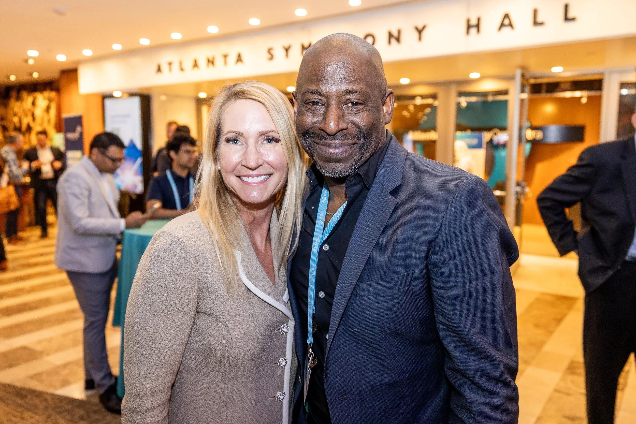 A smiling woman with long blonde hair and a smiling man with a shaved head in a dark suit at the Atlanta Symphony Hall, with other people and event banners in the background.