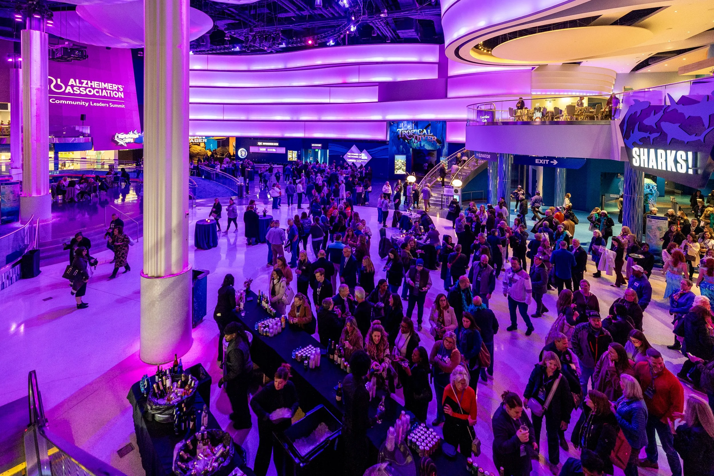 Large indoor event with many people gathered, purple and blue lighting, a bar area with drinks, and signs reading 'Alzheimer's Association', 'Sharks!', and 'Exit'.
