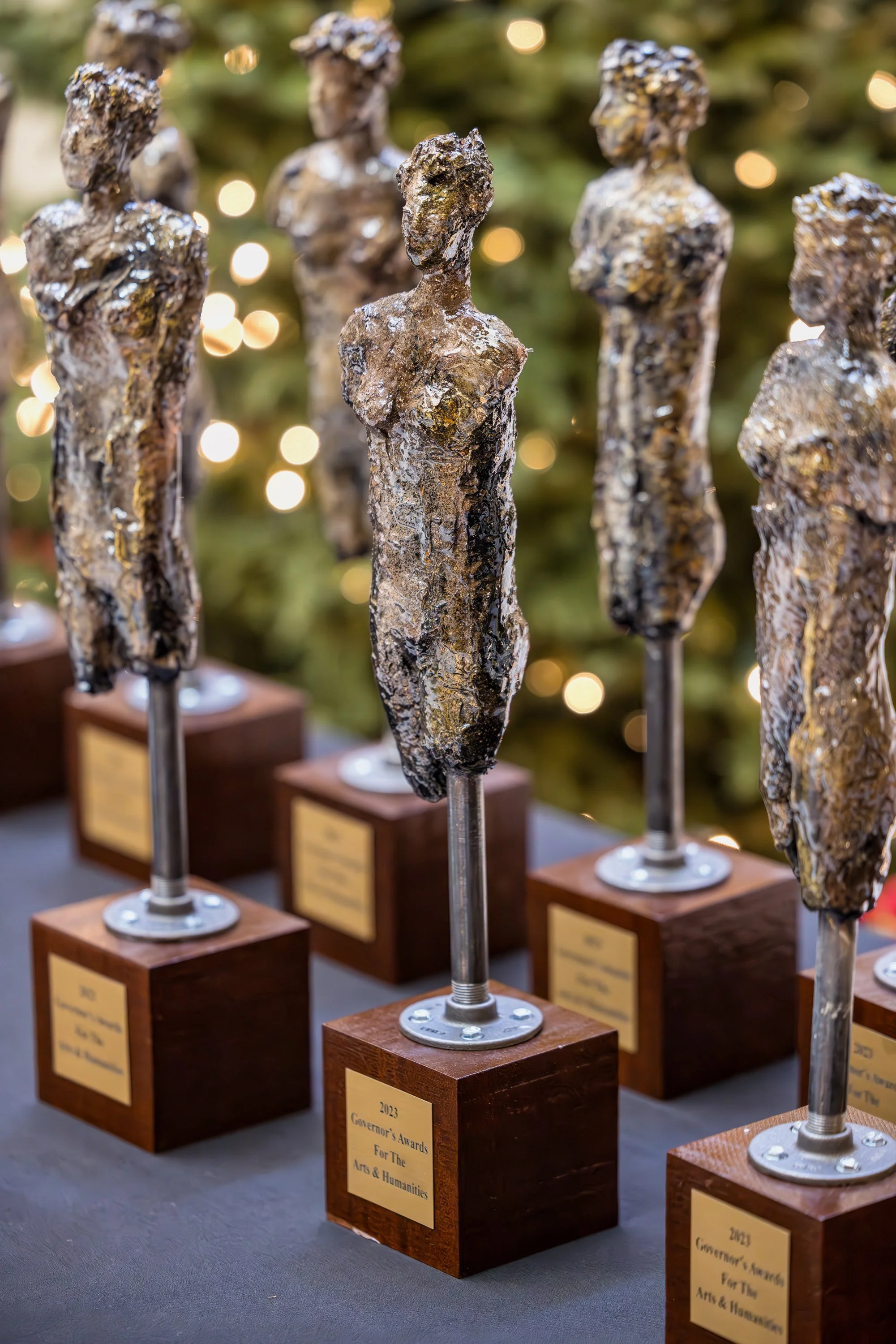Multiple art awards trophies resembling abstract female forms with metallic finishes, mounted on wooden bases with plaques, displayed on a table.